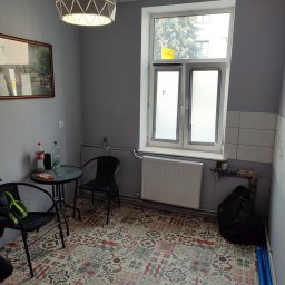 BurbanBud - Pokój po odświeżeniu: szare ściany, wzorzysta podłoga, nowoczesna lampa. Widoczne okno, grzejnik, stół z krzesłami, obraz na ścianie.