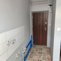 BurbanBud - Korytarz po remoncie: szare ściany, wzorzysta podłoga, brązowe drzwi. Białe płytki z motywem kawy i niebieskie rury. Widoczne piony i poziomy, schludne wykończenie.