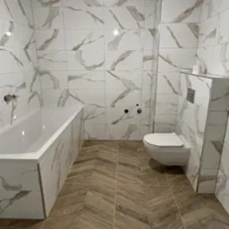 Wykończona łazienka: białe płytki z marmurowym wzorem na ścianach i obudowie wanny, brązowe płytki na podłodze ułożone w jodełkę, podwieszana toaleta.