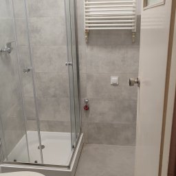 BurbanBud - Wykończona łazienka z kabiną prysznicową, grzejnikiem drabinkowym i toaletą. Szare płytki na ścianach i podłodze. Widoczne detale instalacji i okno z witrażem.