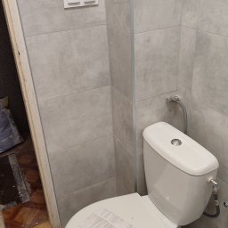 BurbanBud - Jasna łazienka po remoncie: szare płytki na ścianach i podłodze, biała toaleta z dokumentacją na desce, widoczne włączniki światła i fragment odnowionego wejścia.