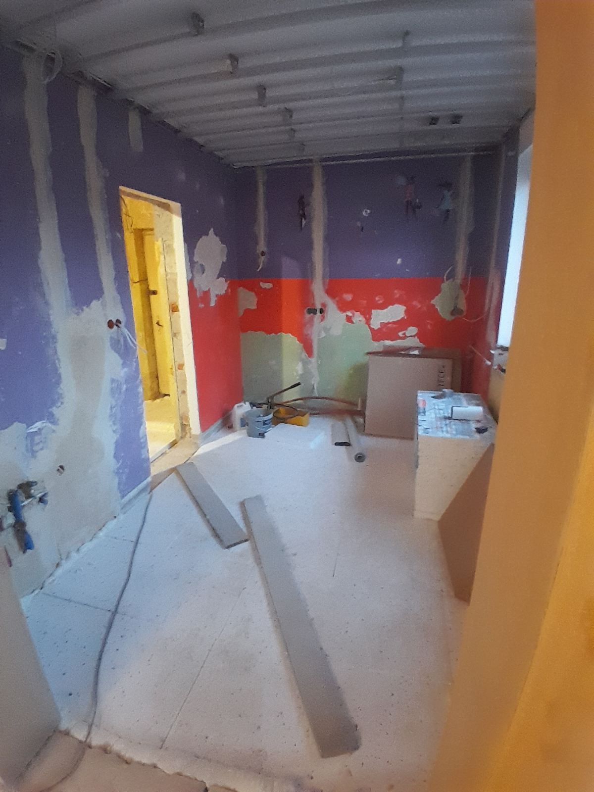 Wnętrze w trakcie remontu: ściany z resztkami farb, surowy sufit, narzędzia i materiały budowlane. Widoczne przygotowania do dalszych prac wykończeniowych.
