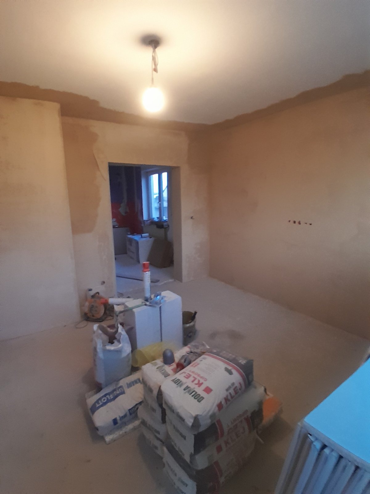 Pokój w trakcie prac wykończeniowych: ściany po gładzi, worki z klejem na podłodze, widoczne narzędzia i materiały budowlane. W tle widoczne okno i inne pomieszczenie.