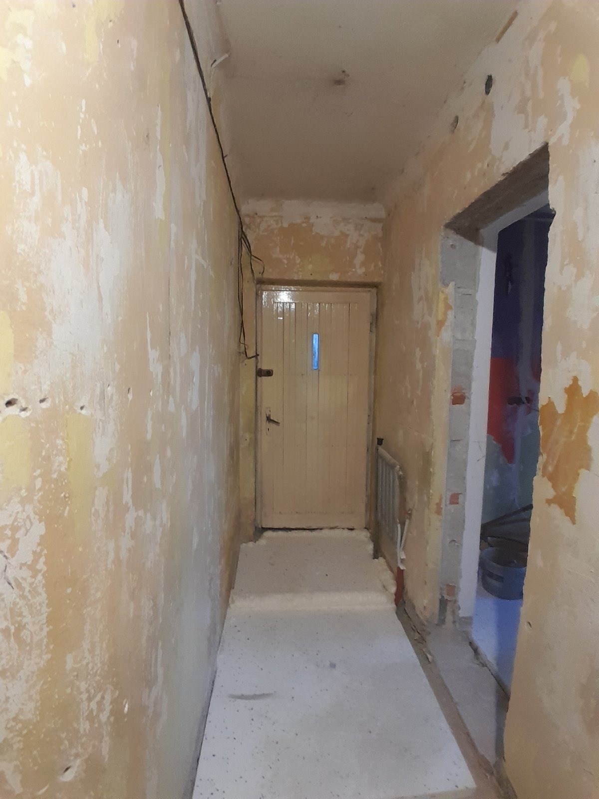 Korytarz w trakcie prac przygotowawczych do remontu. Ściany ze śladami starej farby, widoczne fragmenty instalacji elektrycznej, surowy beton na podłodze.