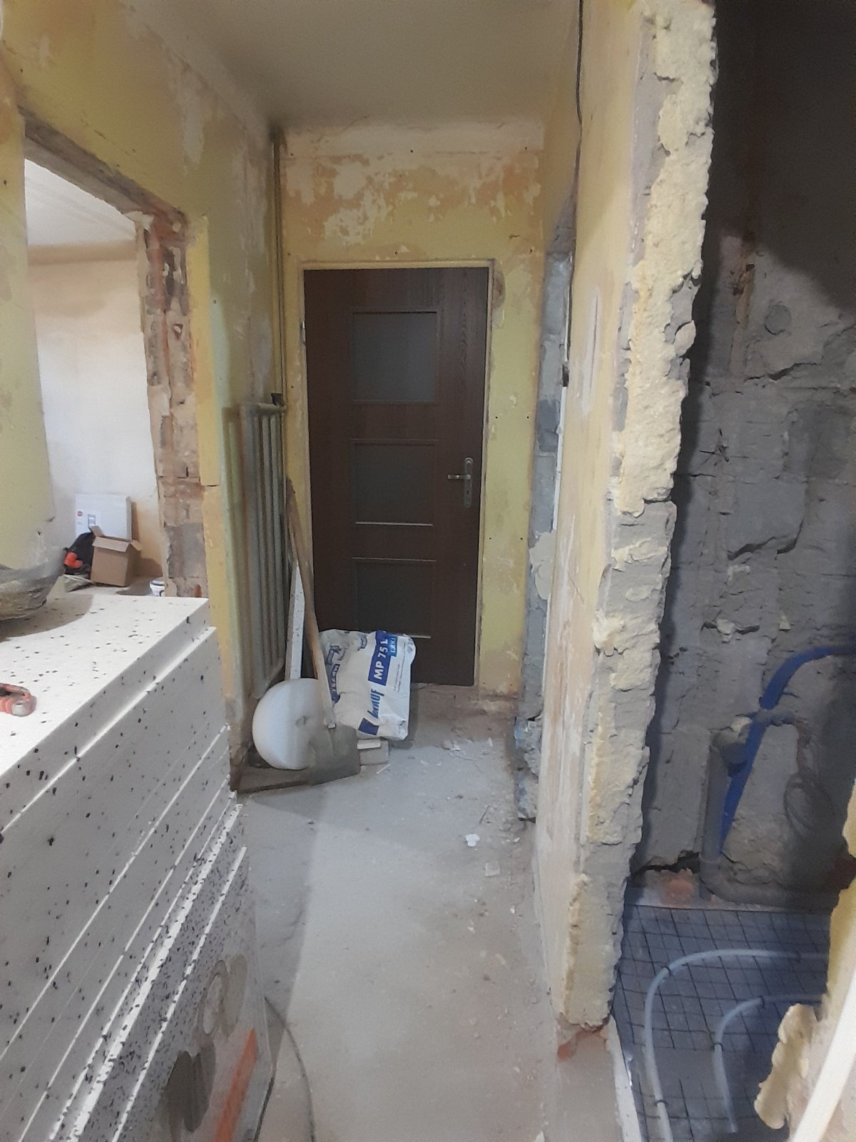 Wnętrze w trakcie remontu: odsłonięte mury, instalacja ogrzewania podłogowego, worki z zaprawą i stos płyt. Widoczne ślady po starych powłokach malarskich.