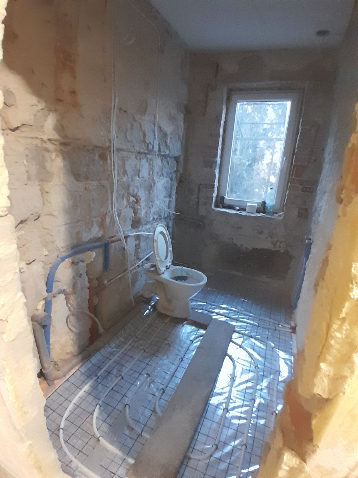 Łazienka w trakcie remontu: surowe ściany, ułożona instalacja ogrzewania podłogowego, widoczna toaleta. Okno z widokiem na zewnątrz.