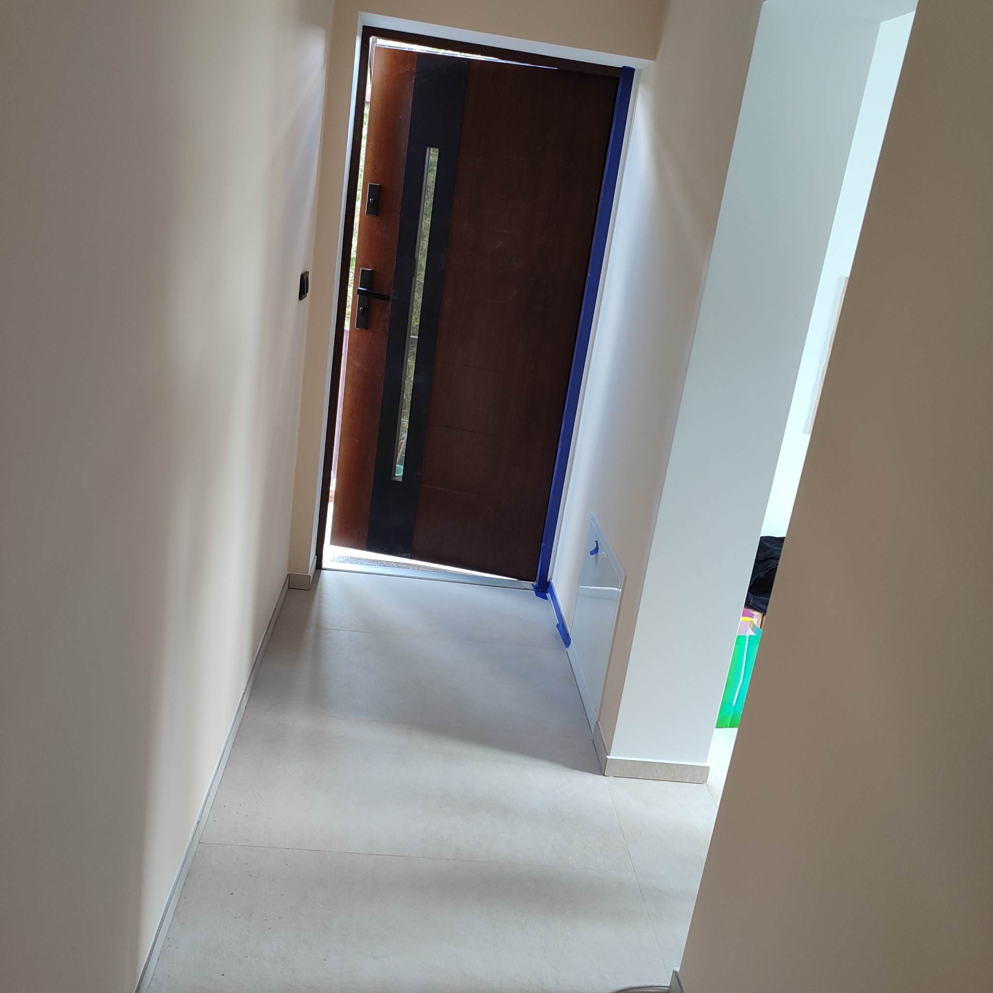 Wnętrze po remoncie: jasne ściany, beżowa podłoga z płytek i ciemne, częściowo otwarte drzwi wejściowe z niebieską taśmą zabezpieczającą.