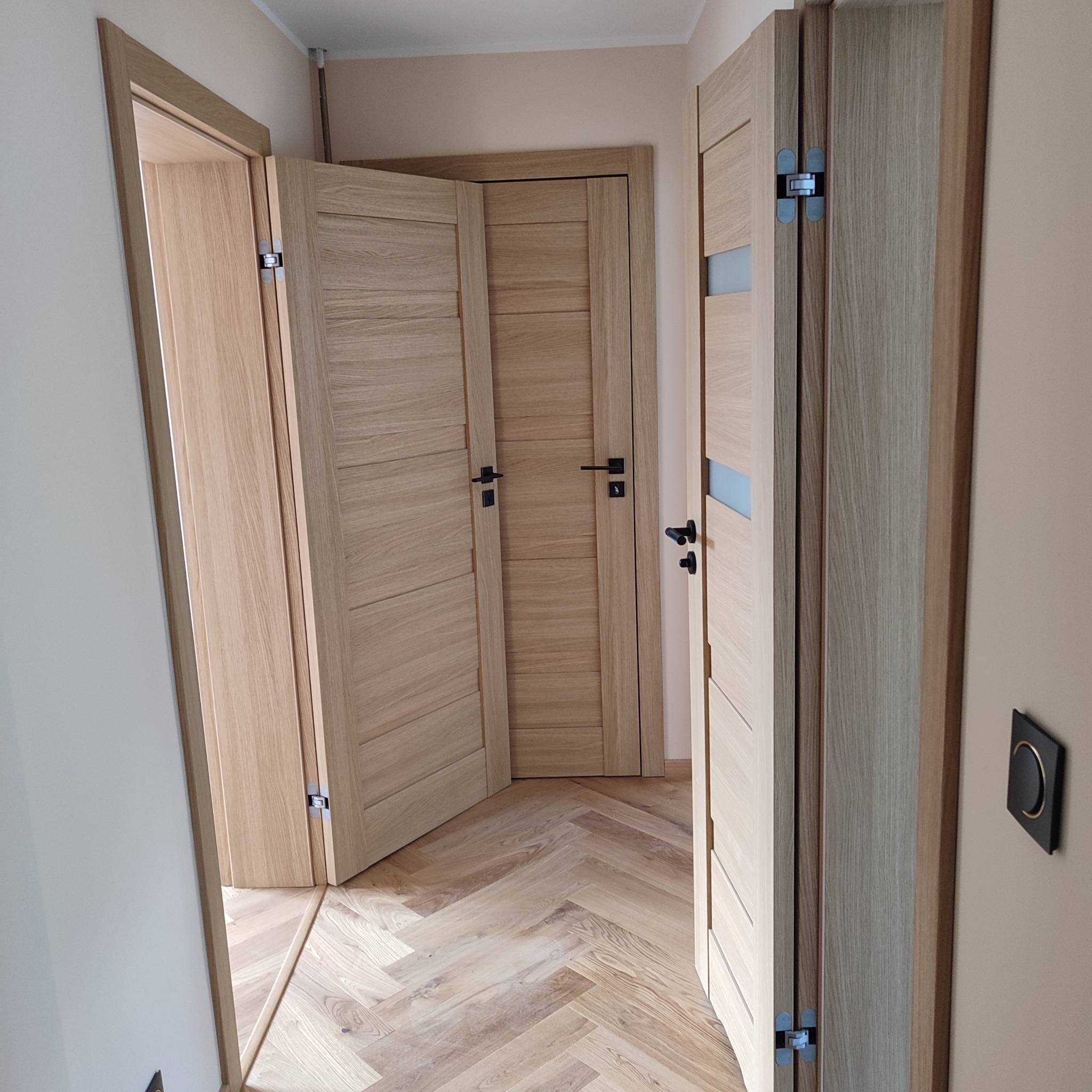 Jasne, drewniane drzwi wewnętrzne zamontowane w mieszkaniu, widoczne zawiasy i klamki. Parkiet ułożony w jodełkę. Minimalistyczny włącznik światła na ścianie.