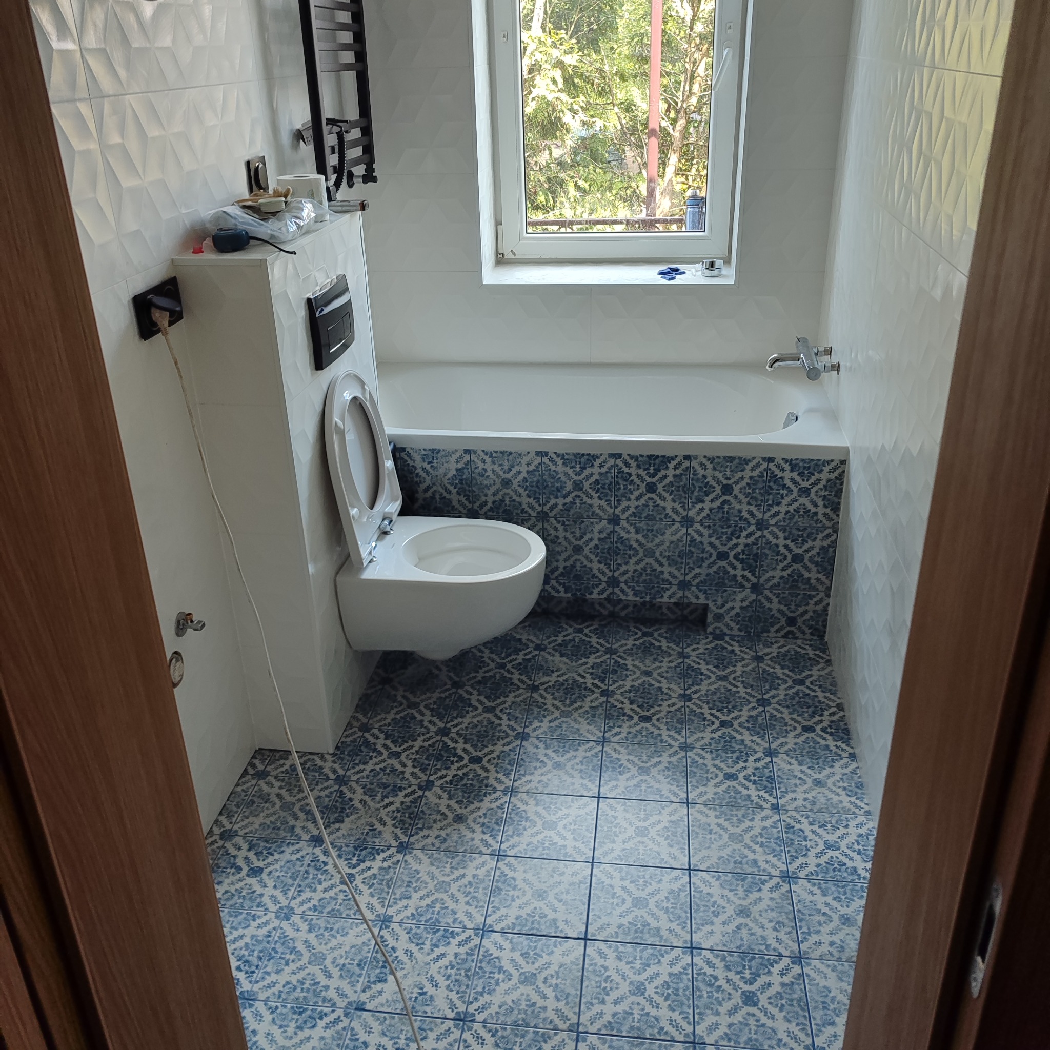 Błękitno-biała łazienka po remoncie: wzorzyste płytki podłogowe i na obudowie wanny, białe płytki 3D na ścianach, wisząca toaleta. Widok na okno.