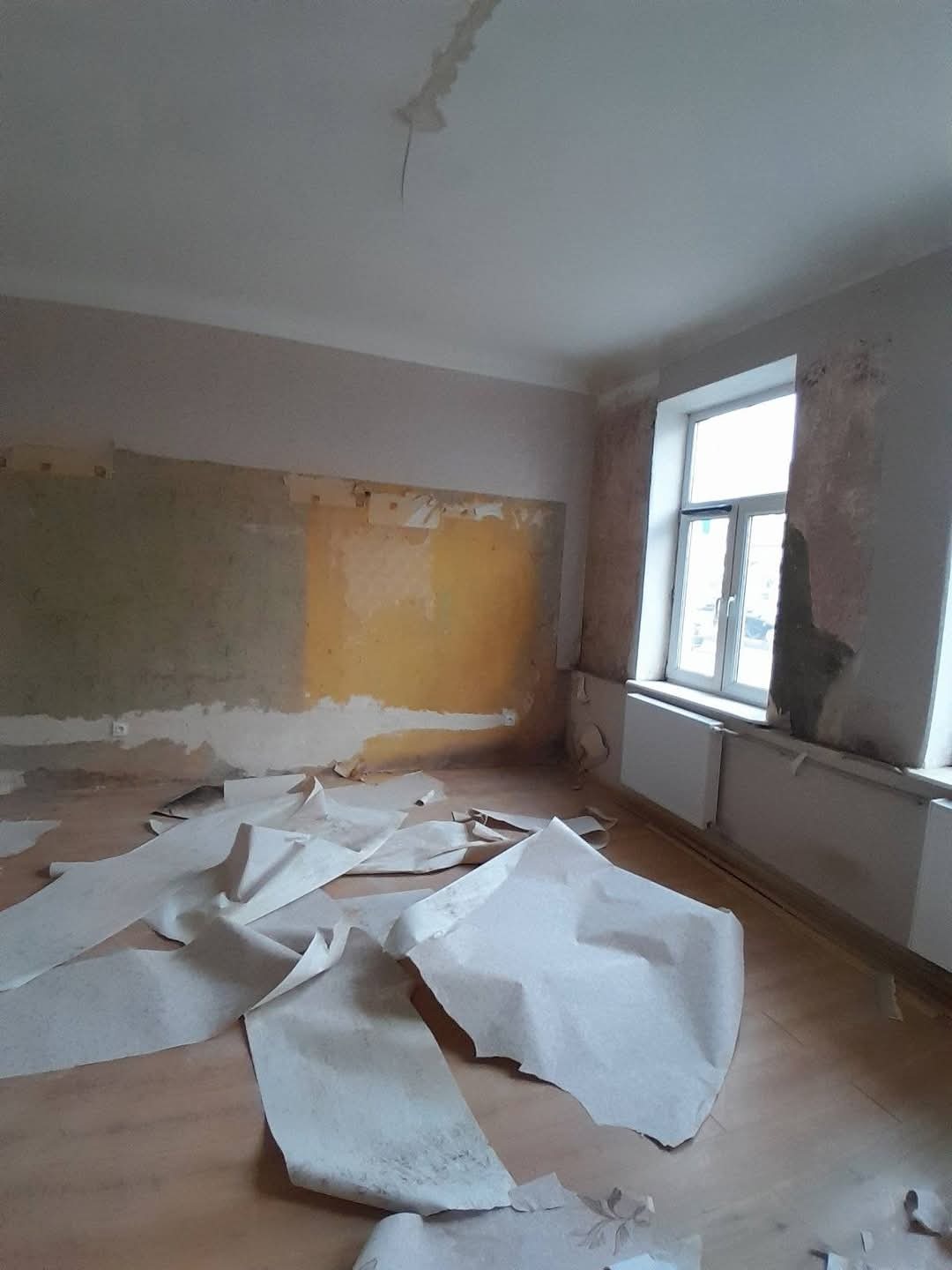 Pokój w trakcie przygotowań do remontu: zerwane tapety, odsłonięte fragmenty ścian, widoczne ślady po farbie i kleju. Okno z grzejnikiem w tle. Bałagan na podłodze.
