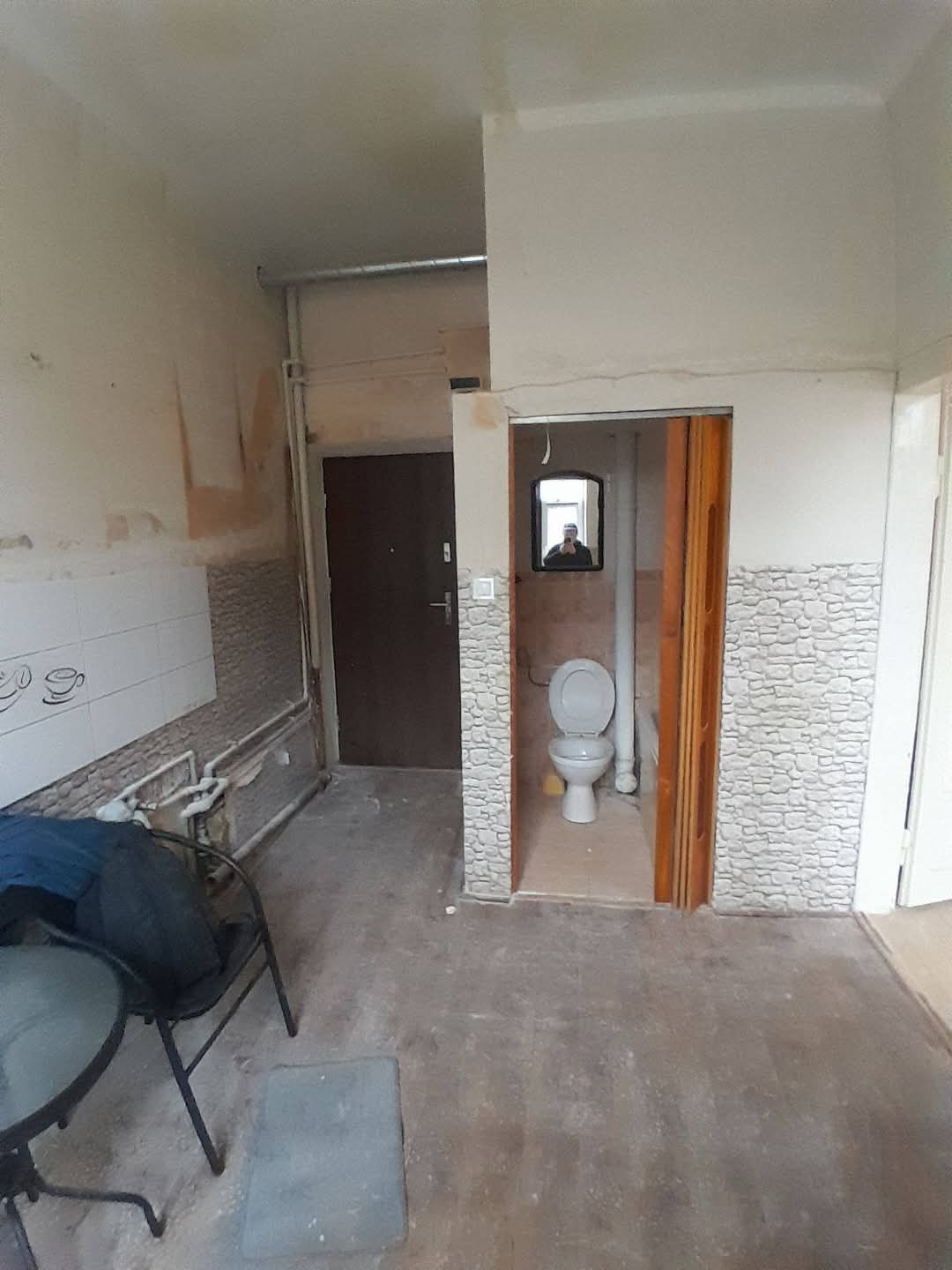 Wnętrze mieszkania w trakcie remontu: widoczne ściany przygotowane do malowania, fragmenty starej glazury, drzwi wejściowe i wejście do toalety z lustrem.