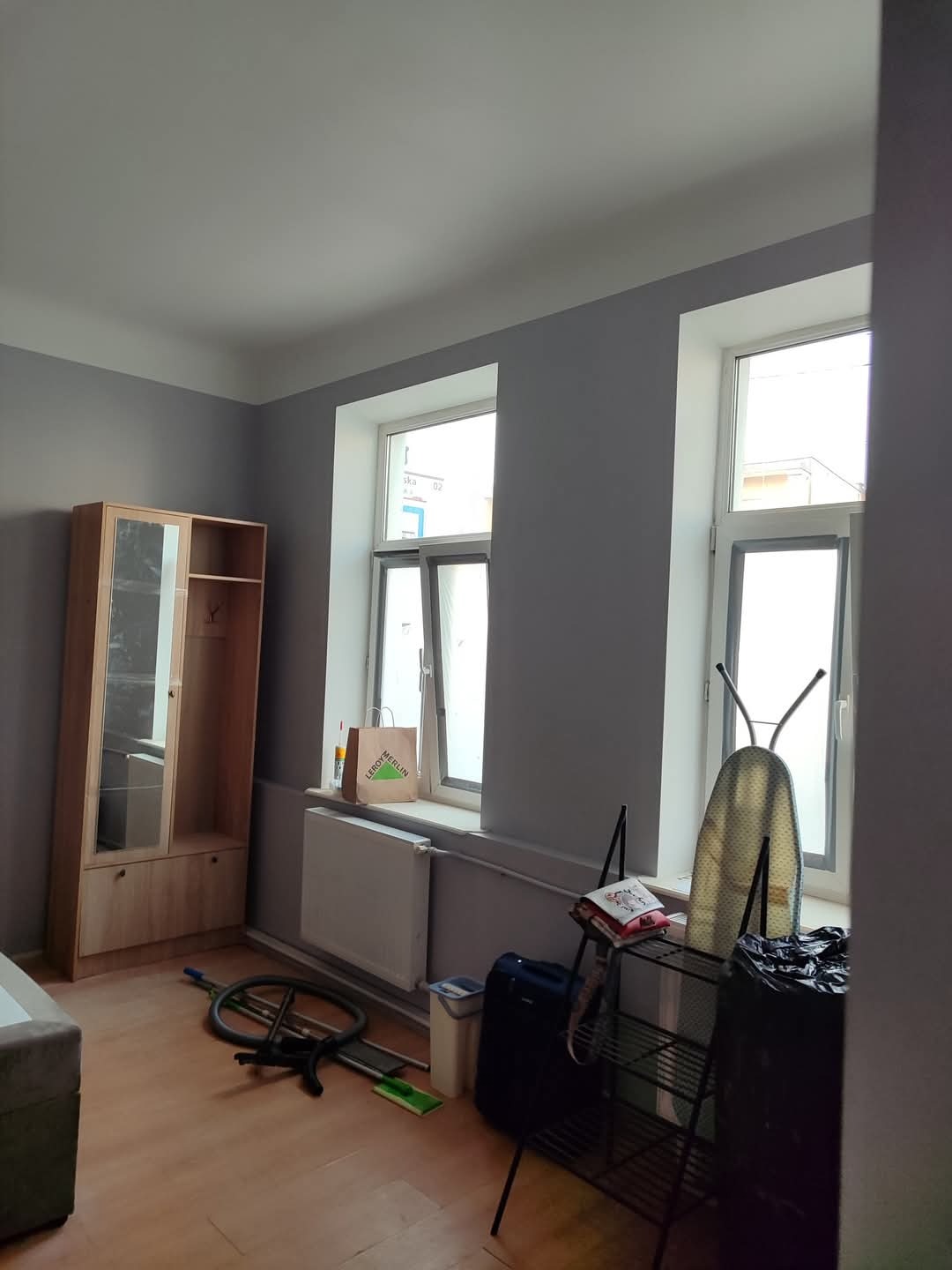 Pomieszczenie po malowaniu ścian na szaro, z widocznym grzejnikiem, oknami, szafką i drobnymi przedmiotami. Widoczne ślady po remoncie.