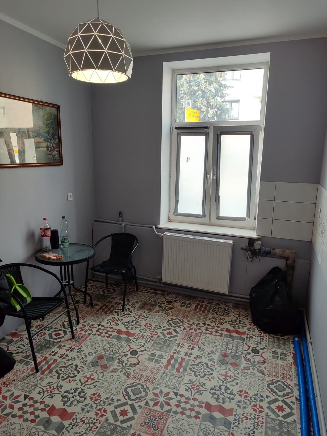 Pokój po odświeżeniu: szare ściany, wzorzysta podłoga, nowoczesna lampa. Widoczne okno, grzejnik, stół z krzesłami, obraz na ścianie.