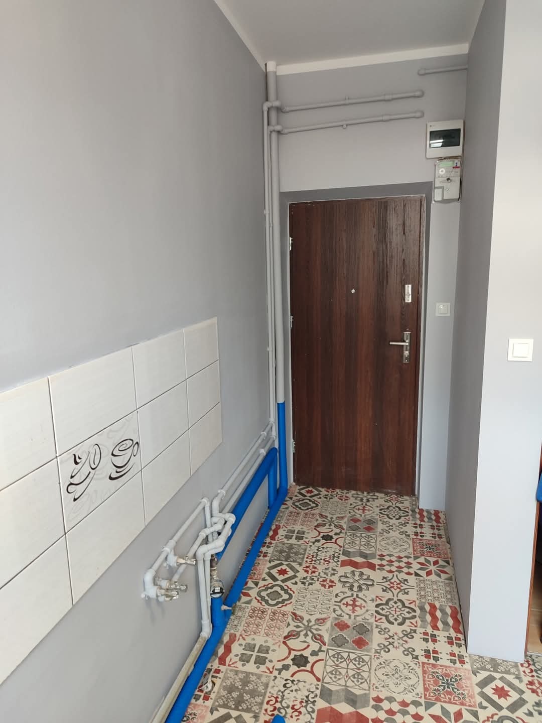 Korytarz po remoncie: szare ściany, wzorzysta podłoga, brązowe drzwi. Białe płytki z motywem kawy i niebieskie rury. Widoczne piony i poziomy, schludne wykończenie.