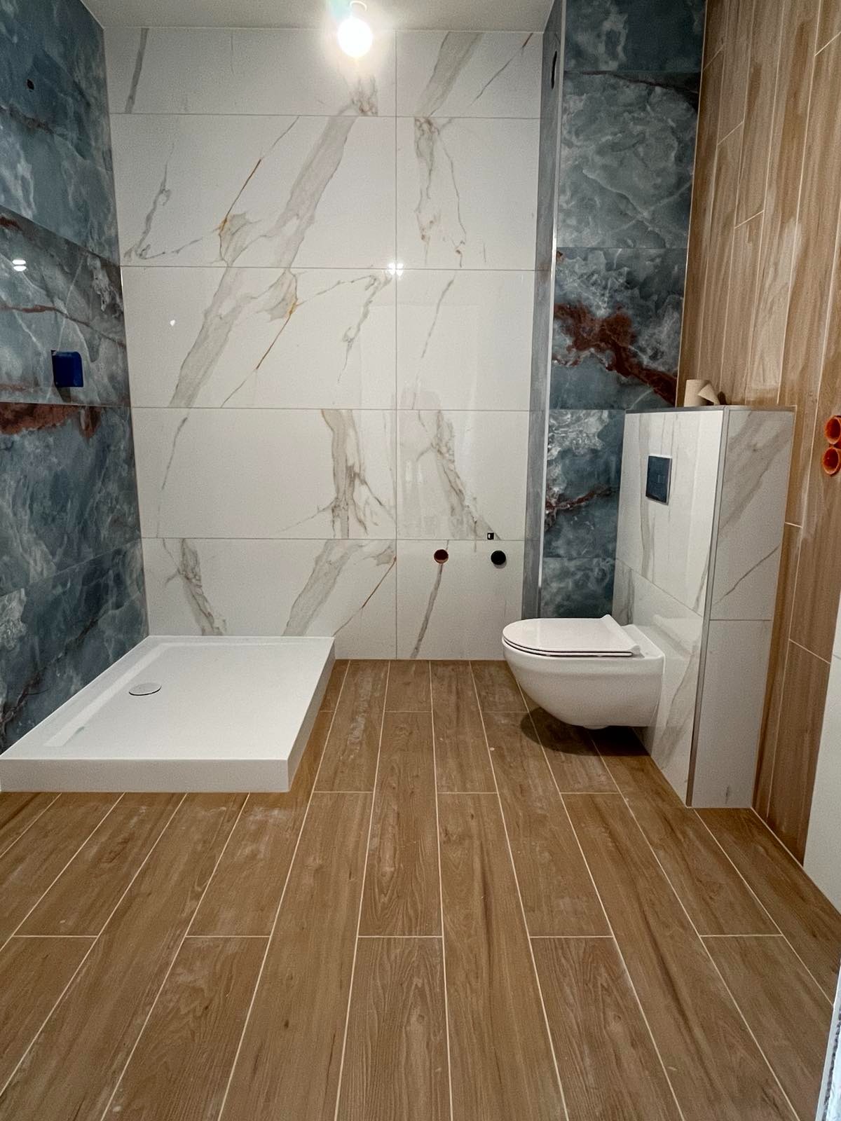 Nowoczesna łazienka po remoncie: kabina prysznicowa, podwieszana toaleta, marmurowe i drewnopodobne płytki. Całość w jasnych barwach.