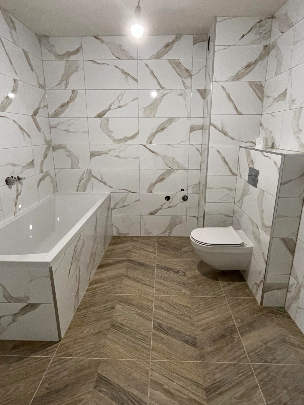 Wykończona łazienka: białe płytki z marmurowym wzorem na ścianach i obudowie wanny, brązowe płytki na podłodze ułożone w jodełkę, podwieszana toaleta.