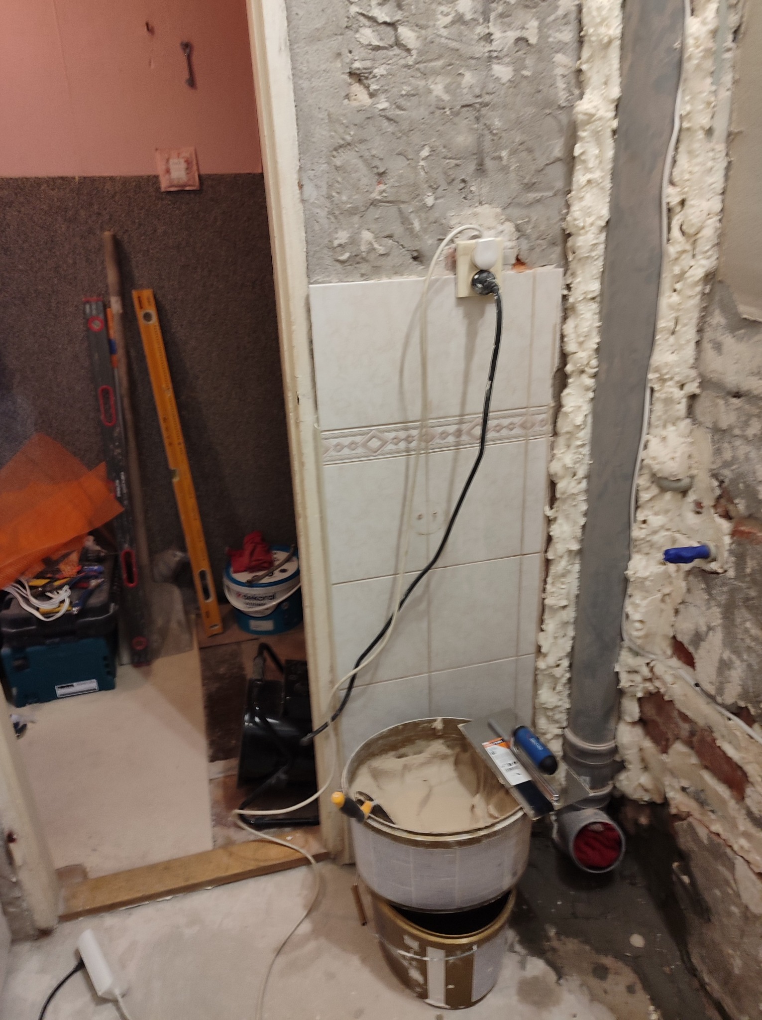 Niedokończona łazienka w trakcie prac: widoczne płytki, puszka z zaprawą, narzędzia glazurnicze, rury i pianka montażowa. Fragment ściany bez tynku.