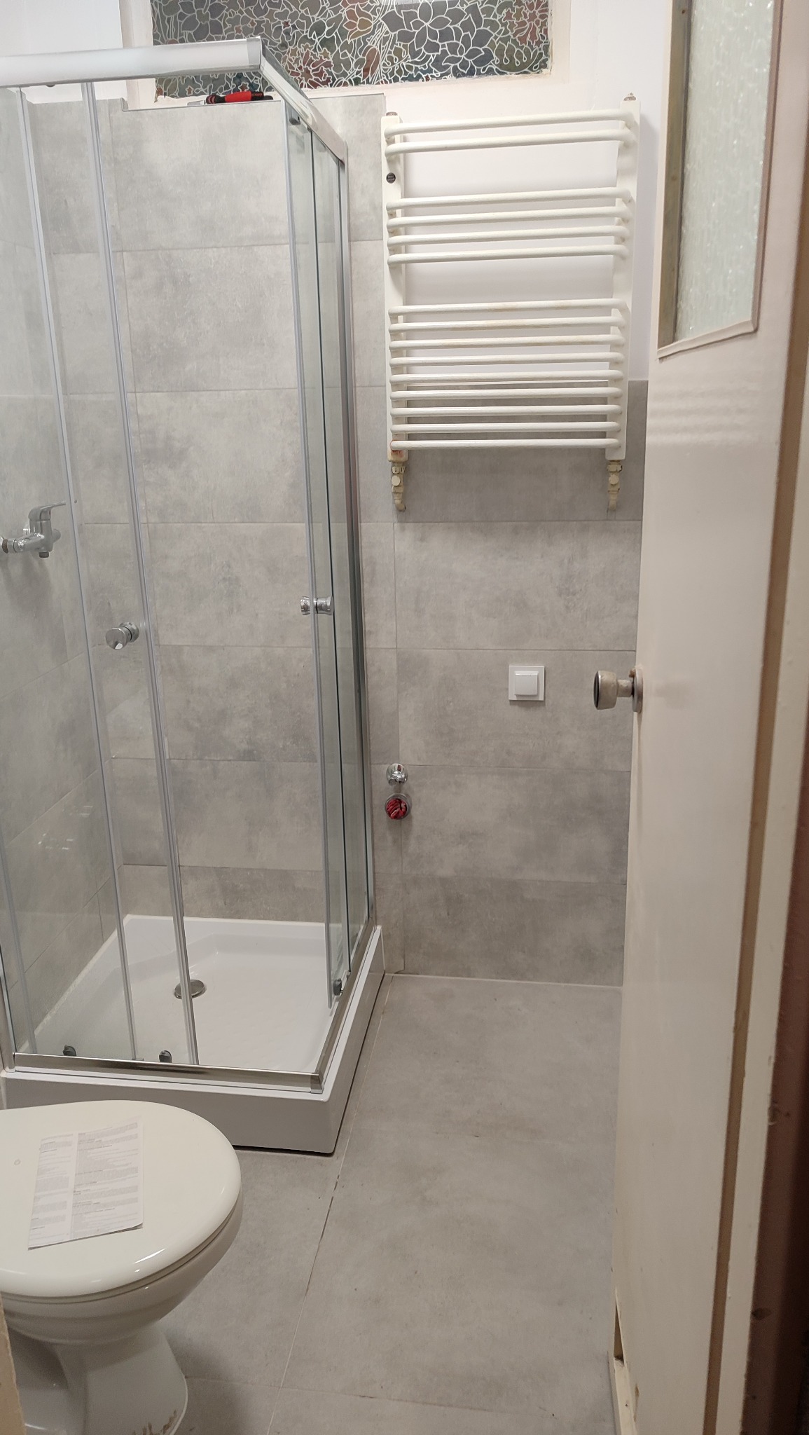 Wykończona łazienka z kabiną prysznicową, grzejnikiem drabinkowym i toaletą. Szare płytki na ścianach i podłodze. Widoczne detale instalacji i okno z witrażem.