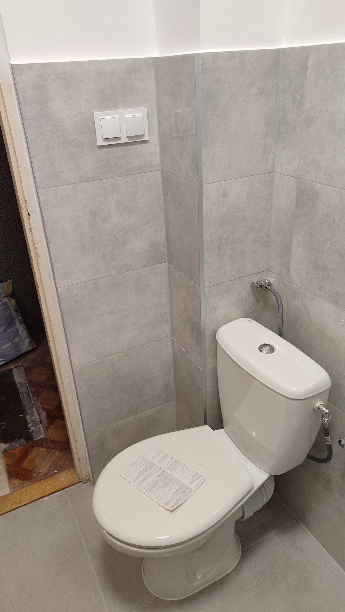 Jasna łazienka po remoncie: szare płytki na ścianach i podłodze, biała toaleta z dokumentacją na desce, widoczne włączniki światła i fragment odnowionego wejścia.