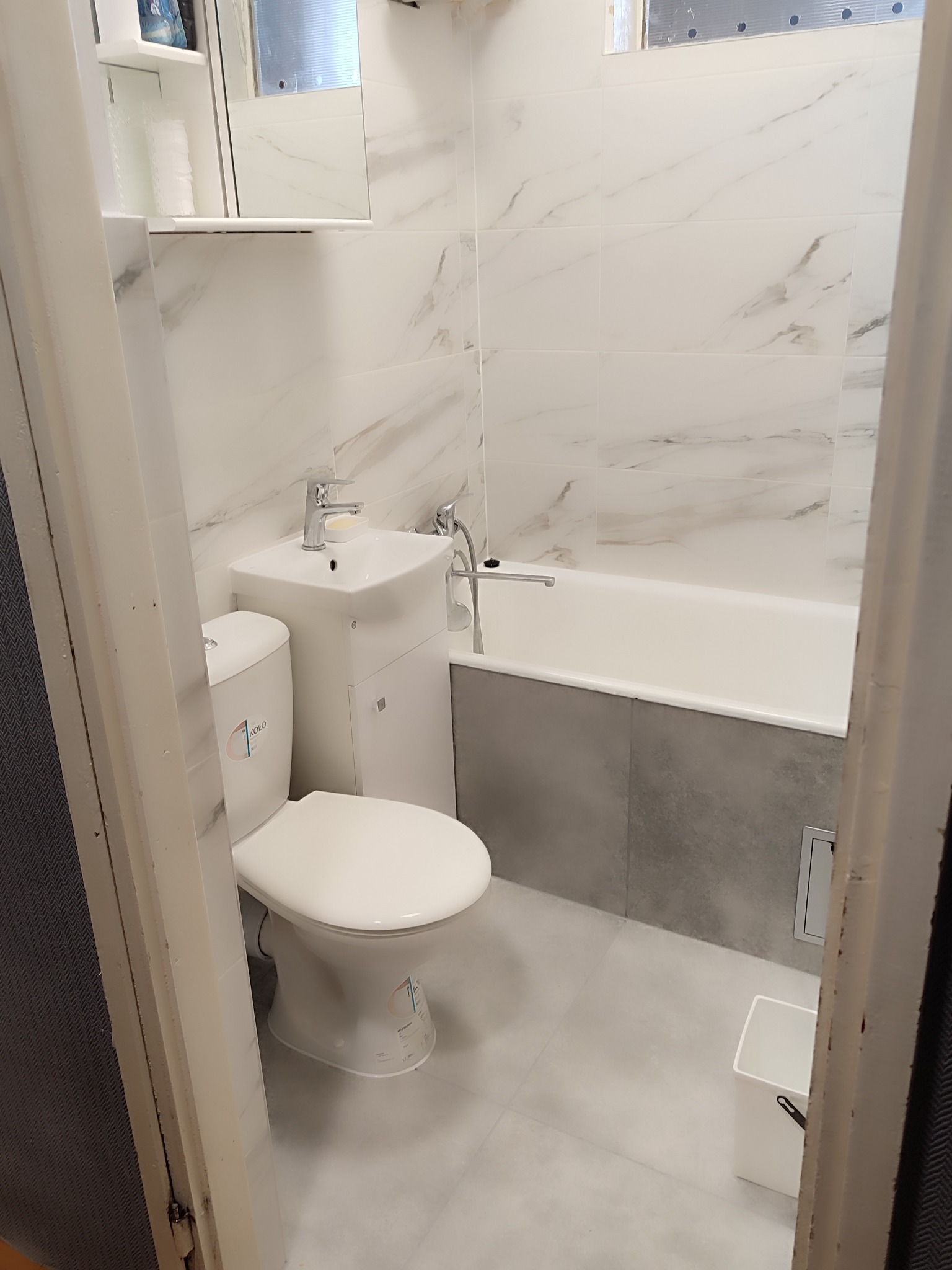 Jasna łazienka po remoncie: białe płytki imitujące marmur, umywalka z szafką, toaleta marki Koło i wanna obudowana szarą mozaiką. Widok zza futryny.