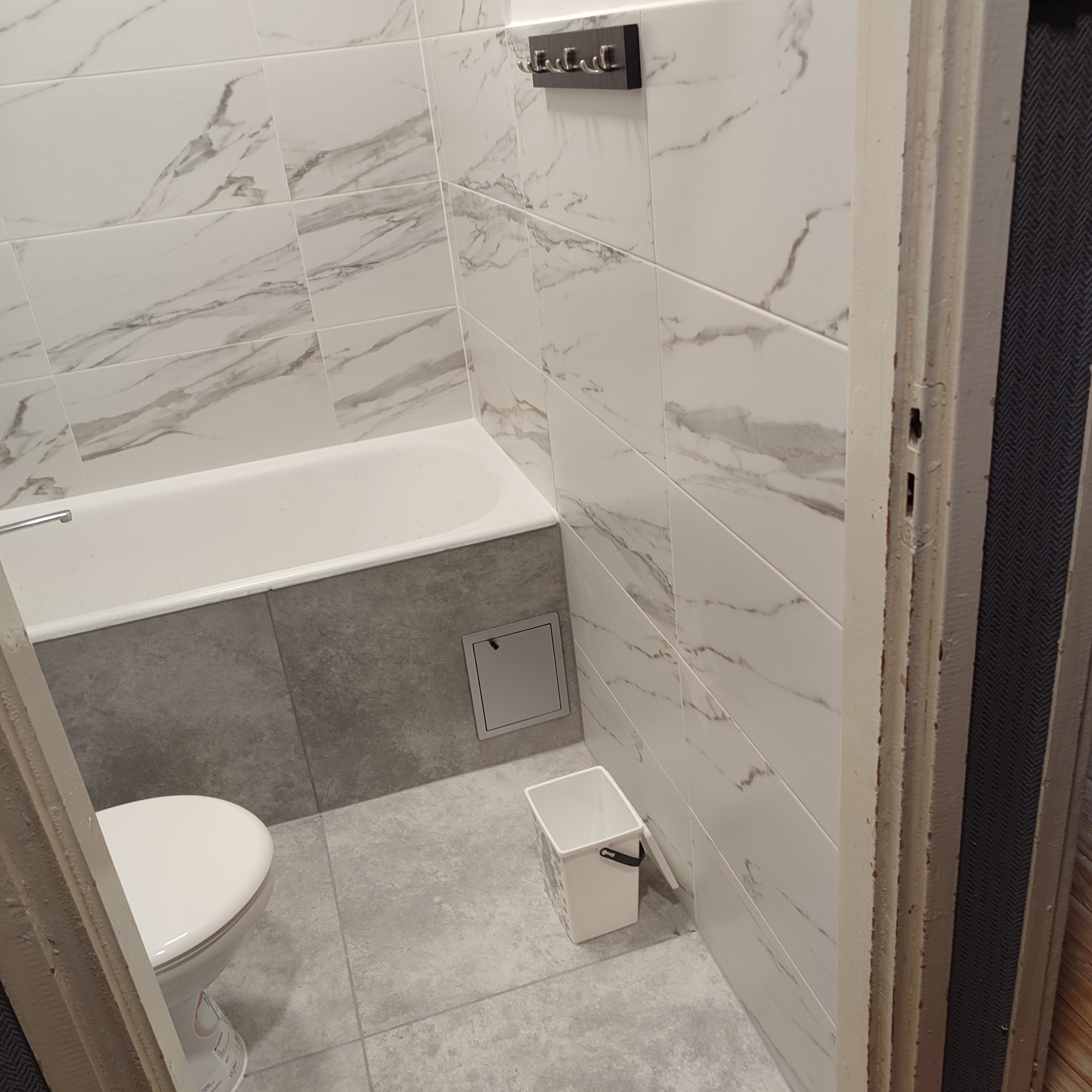 Wykończona łazienka z wanną obudowaną szarą płytką, białą marmurową glazurą na ścianach i szarą podłogą. Widoczny fragment toalety i kosz na śmieci.