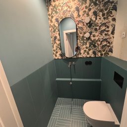 Maestro - Turkusowa łazienka z wzorzystą tapetą i owalnym lustrem. Umywalka w zabudowie z baterią i podwieszana toaleta. Płytki podłogowe ułożone w jodełkę. Nowoczesny design.