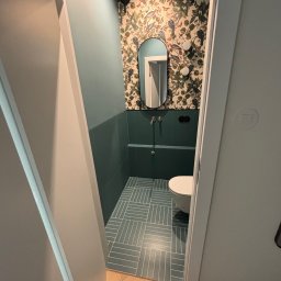 Maestro - Wykończona łazienka z zielonymi płytkami ułożonymi w jodełkę, ozdobną tapetą w rośliny i owalne lustro. Widok z korytarza przez otwarte drzwi.