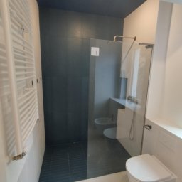 Maestro - Nowoczesna łazienka z kabiną prysznicową typu walk-in, ciemnymi płytkami i białym grzejnikiem drabinkowym. Widoczne elementy armatury i toaleta w minimalistycznym stylu.