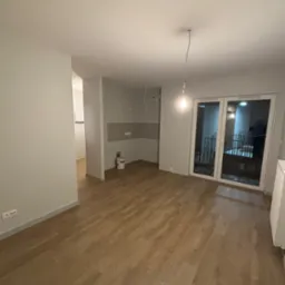 Jasne wnętrze po remoncie: parkiet, świeżo pomalowane ściany w odcieniach szarości, duże okno balkonowe. Widoczny fragment instalacji elektrycznej.