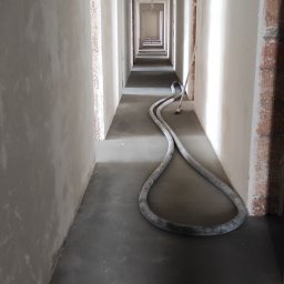 Maestro - Długi korytarz w stanie surowym po nałożeniu tynków, widoczny wąż do tynkowania leżący na podłodze, ściany o nierównej powierzchni przygotowane do dalszych prac wykończeniowych.