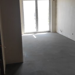 Maestro - Surowe wnętrze z wylewką i gładkimi ścianami przed malowaniem, widoczne przewody elektryczne zwisające z sufitu i duże okno balkonowe wpuszczające jasne światło.