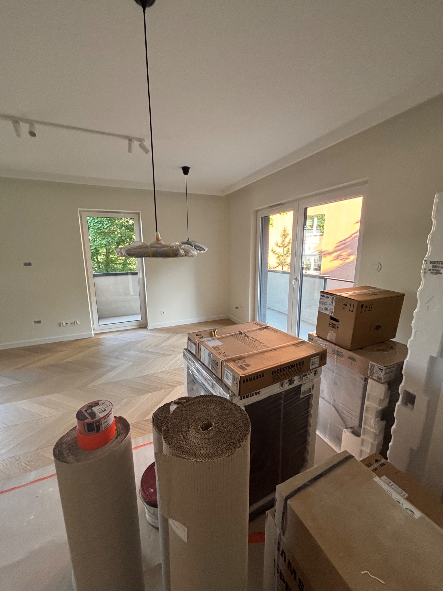 Jasne wnętrze w trakcie wykańczania: parkiet w jodełkę, lampy wiszące w folii ochronnej, kartony z nowym sprzętem AGD. Widok na balkon i zieleń.
