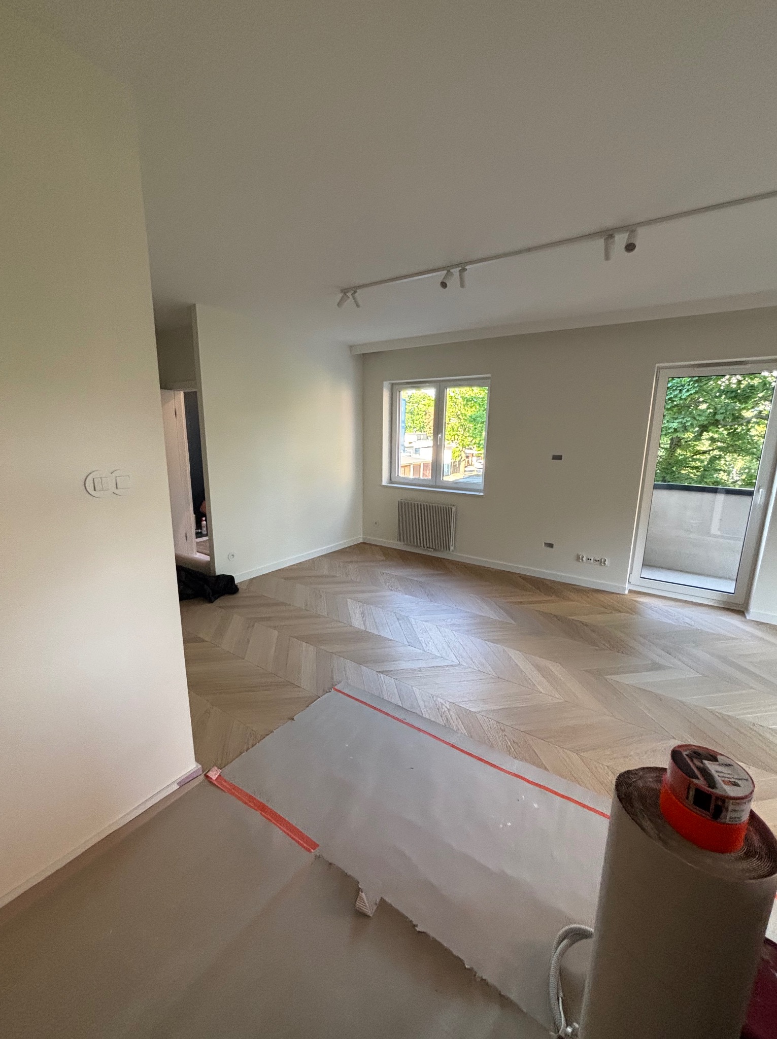 Parkiet w jodełkę francuską w jasnym, przestronnym pokoju z oknem i wyjściem na balkon. Część podłogi zabezpieczona folią malarską.