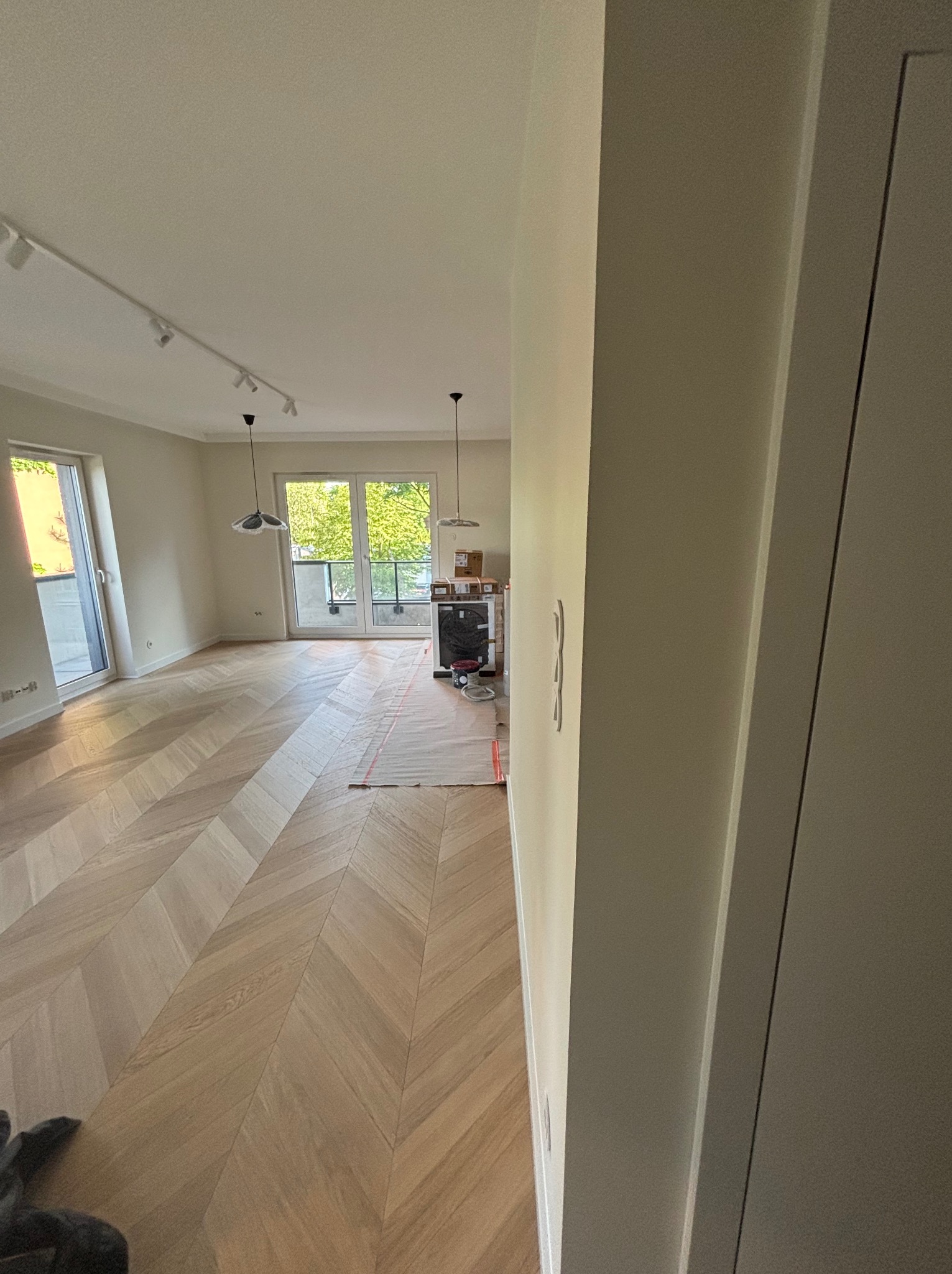 Jasne, nowo położone parkiety w jodełkę dominują w przestronnym wnętrzu z balkonem. Widoczne lampy sufitowe i przygotowania do dalszych prac wykończeniowych.