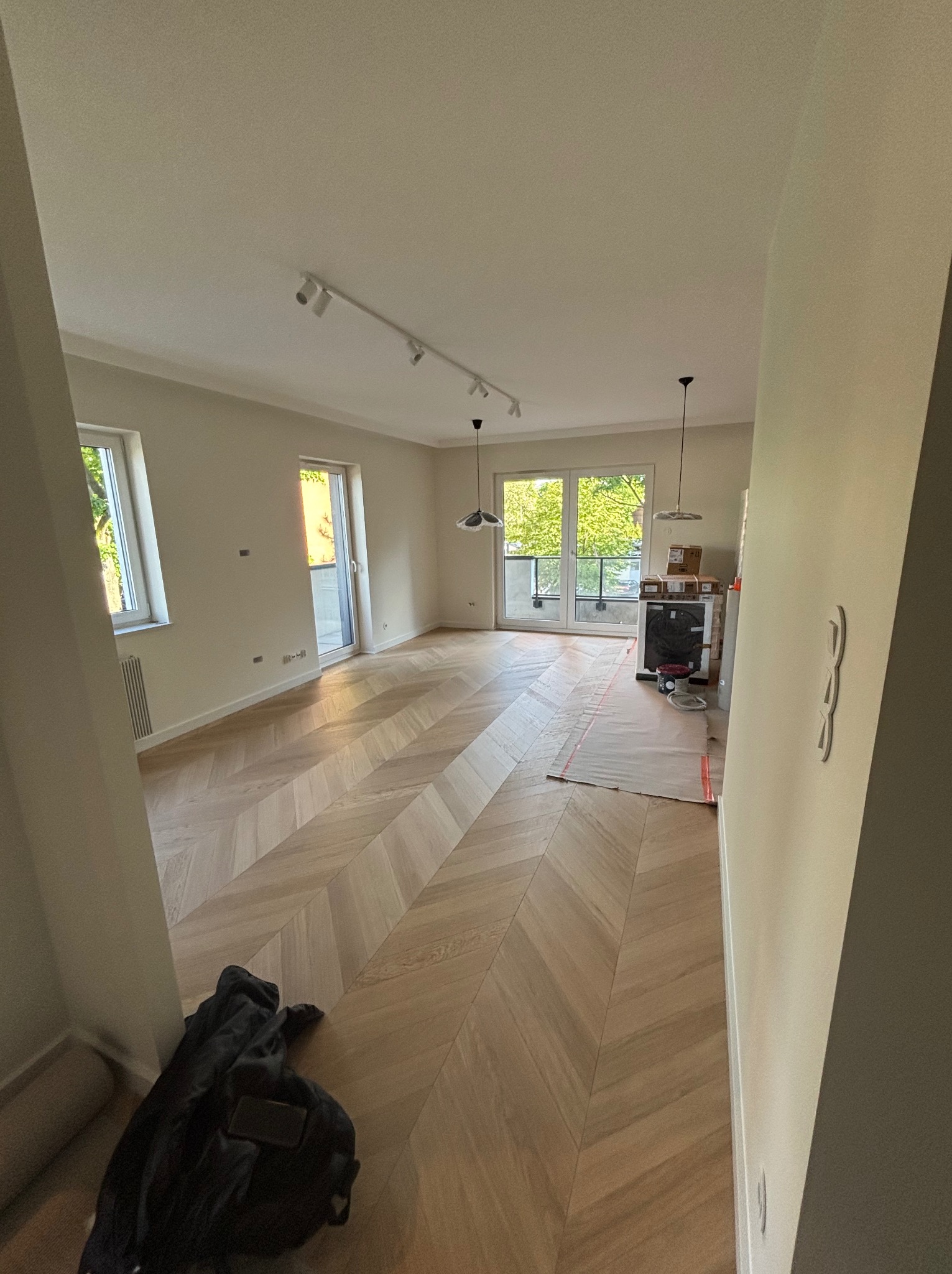 Jasne, przestronne wnętrze po remoncie z parkietem w jodełkę, białymi ścianami i nowoczesnym oświetleniem. Częściowo zabezpieczona podłoga wskazuje na trwające prace.