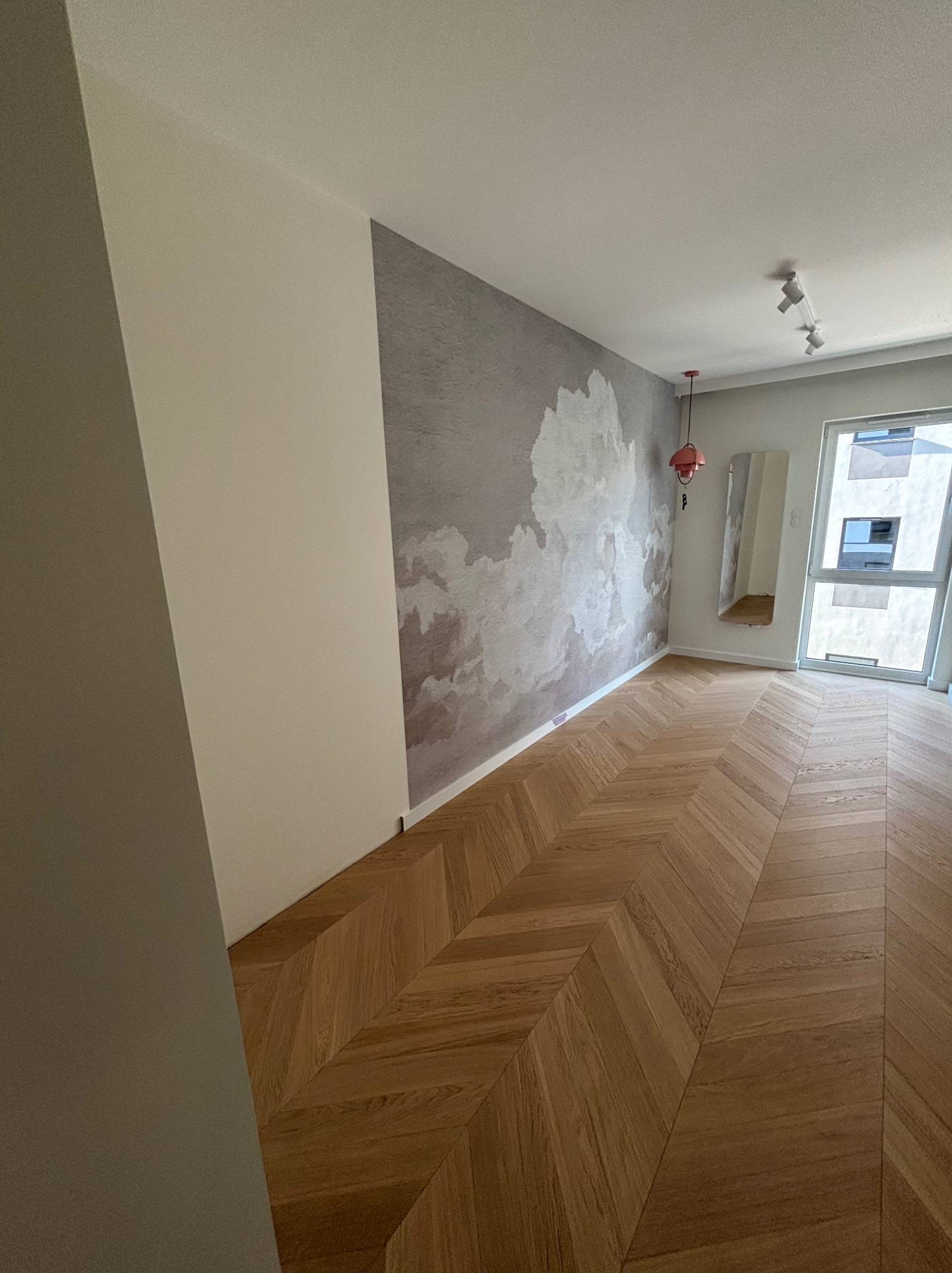 Pokój po remoncie: parkiet w jodełkę, ściana z efektem chmur, minimalistyczna lampa i lustro. Widok na okno z nowym budynkiem w tle.