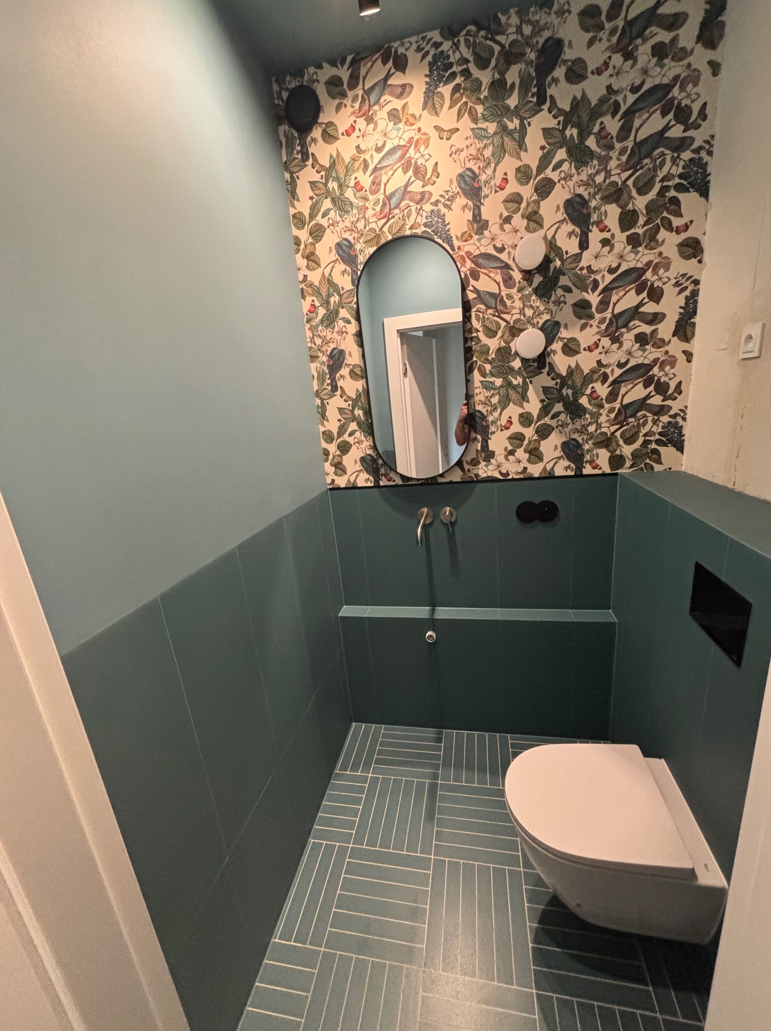 Turkusowa łazienka z wzorzystą tapetą i owalnym lustrem. Umywalka w zabudowie z baterią i podwieszana toaleta. Płytki podłogowe ułożone w jodełkę. Nowoczesny design.