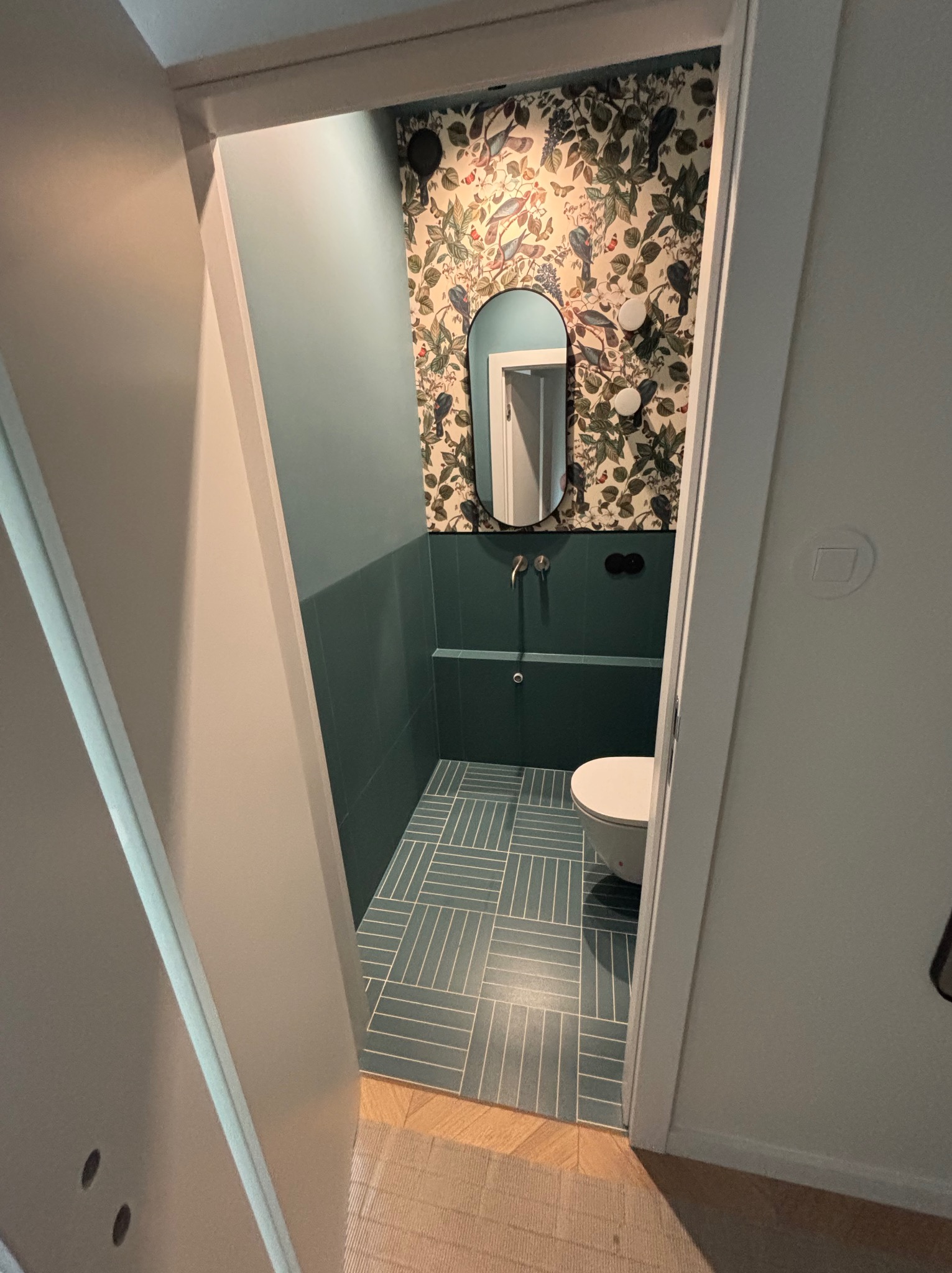 Wykończona łazienka z zielonymi płytkami ułożonymi w jodełkę, ozdobną tapetą w rośliny i owalne lustro. Widok z korytarza przez otwarte drzwi.