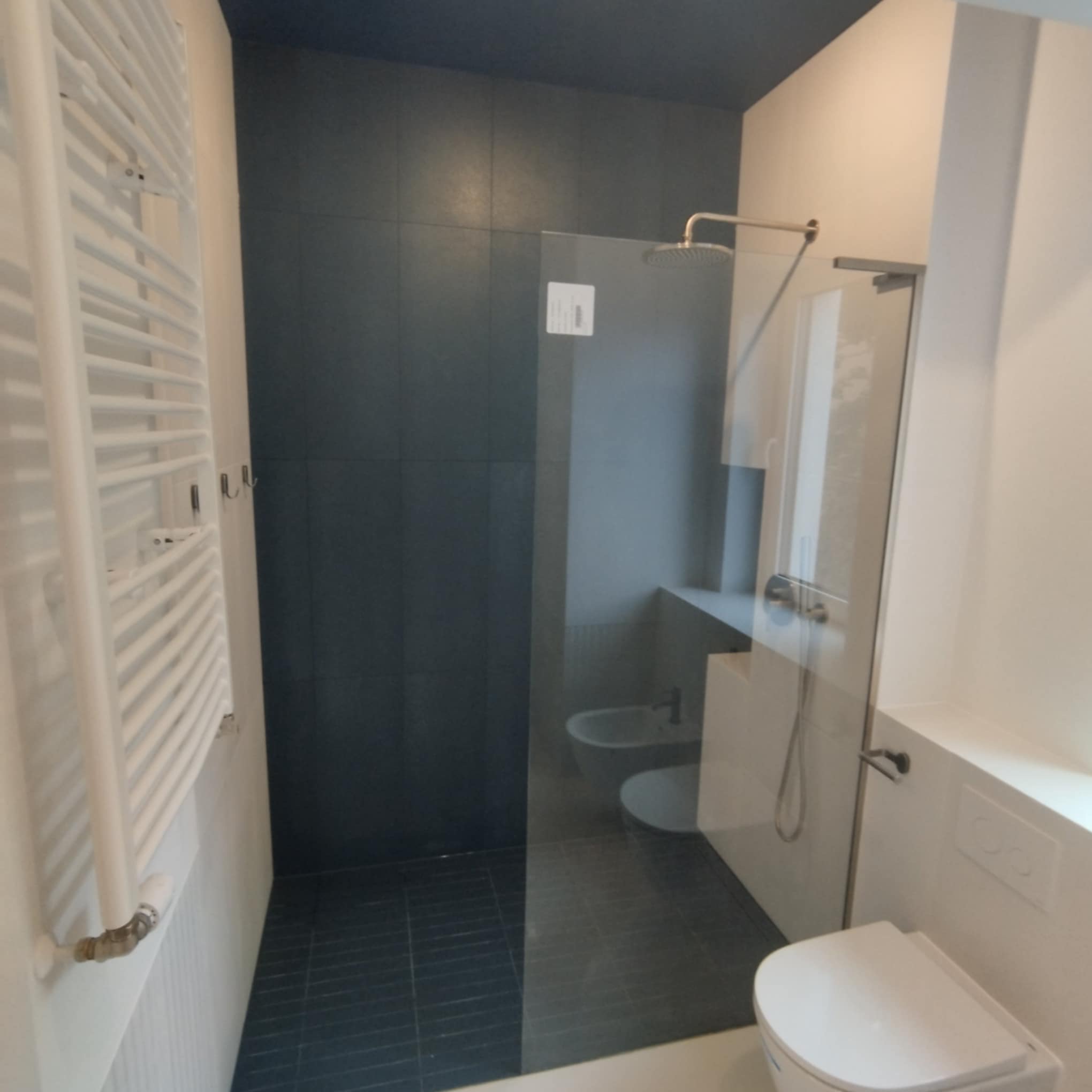 Nowoczesna łazienka z kabiną prysznicową typu walk-in, ciemnymi płytkami i białym grzejnikiem drabinkowym. Widoczne elementy armatury i toaleta w minimalistycznym stylu.