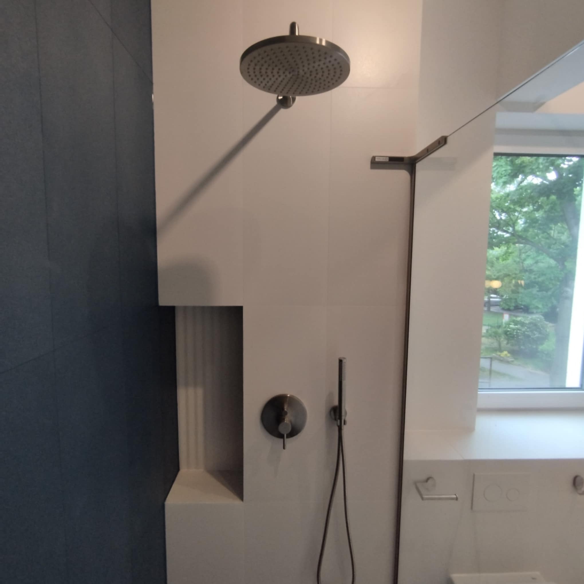 Nowoczesna kabina prysznicowa z deszczownicą i wnęką w ścianie wyłożonej białymi płytkami, z elementami armatury w kolorze grafitowym.