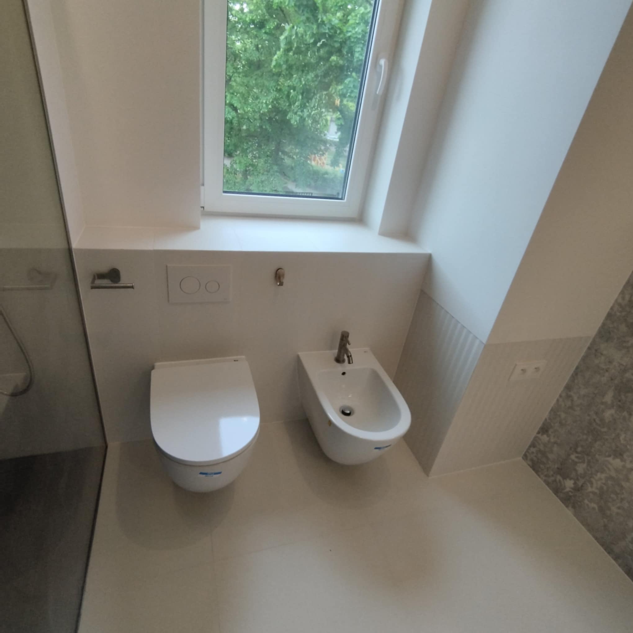 Nowoczesna, jasna łazienka po remoncie. Wisząca toaleta i bidet obok siebie, nad nimi okno z widokiem na zieleń. Ściany i podłoga w odcieniach bieli i szarości.