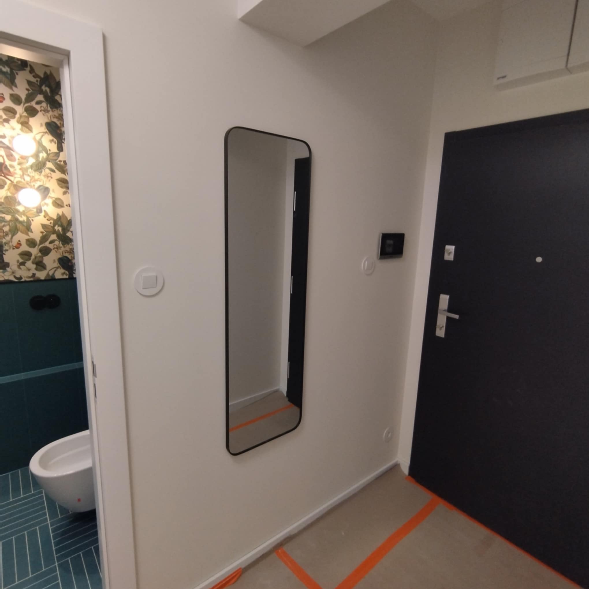 Nowoczesne wykończenie wnętrza: minimalistyczne lustro w czarnej ramie, drzwi wejściowe i fragment łazienki z wzorzystą tapetą i bidetem.