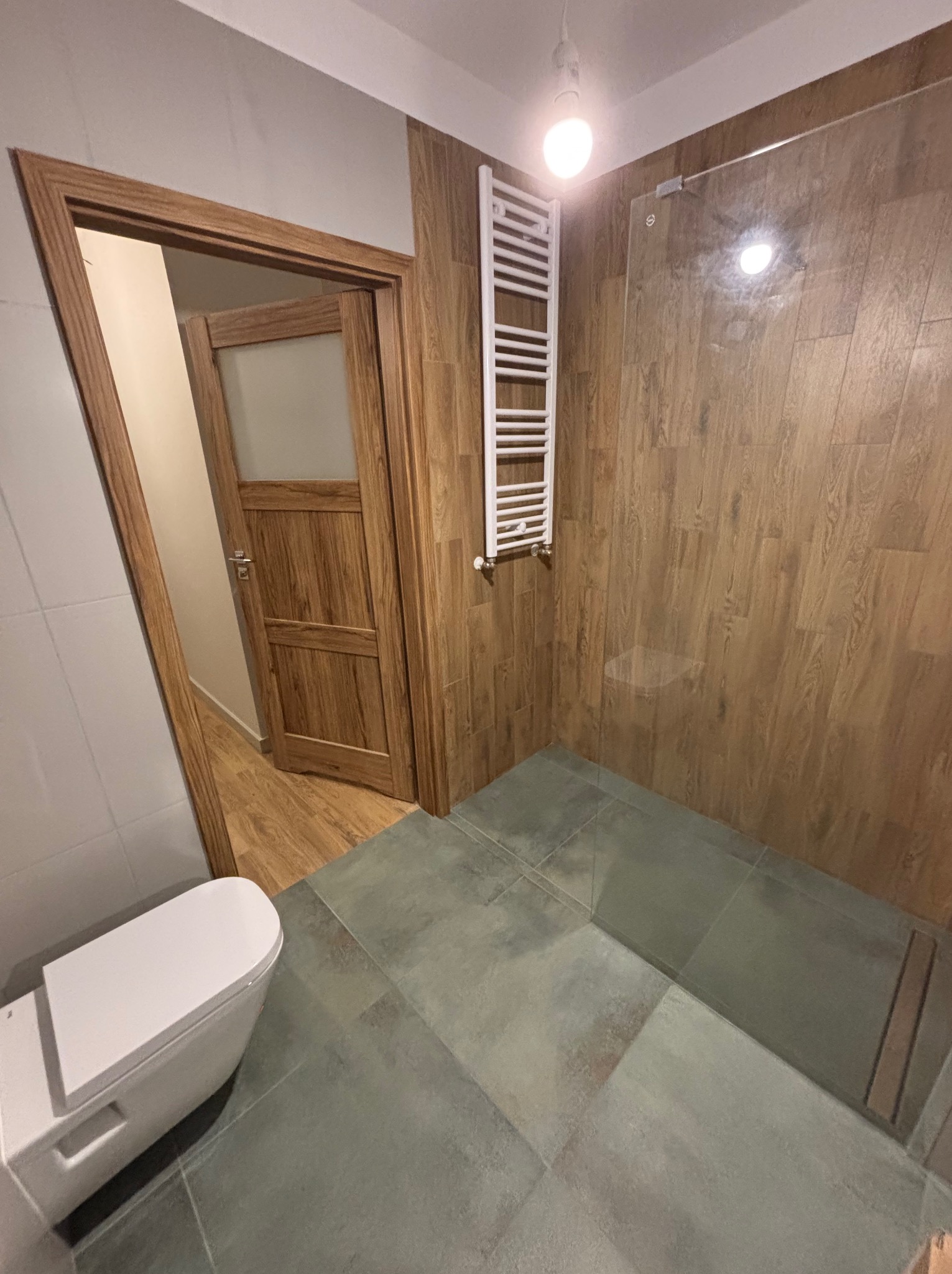 Wykończona łazienka z kabiną prysznicową typu walk-in, drewnopodobnymi płytkami na ścianach i grzejnikiem drabinkowym. Widoczny fragment toalety i otwarte drewniane drzwi.