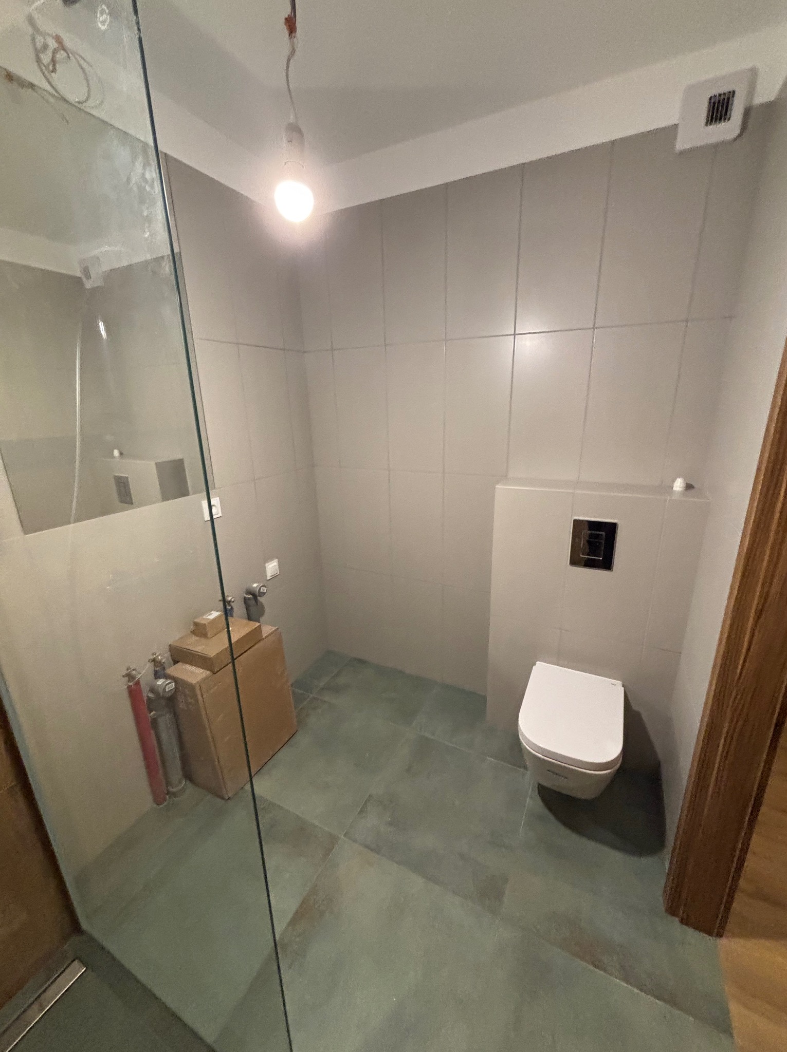 Wykończona łazienka z podwieszaną toaletą i szklaną kabiną prysznicową. Widoczne płytki w odcieniach szarości i elementy instalacji przed zabudową.