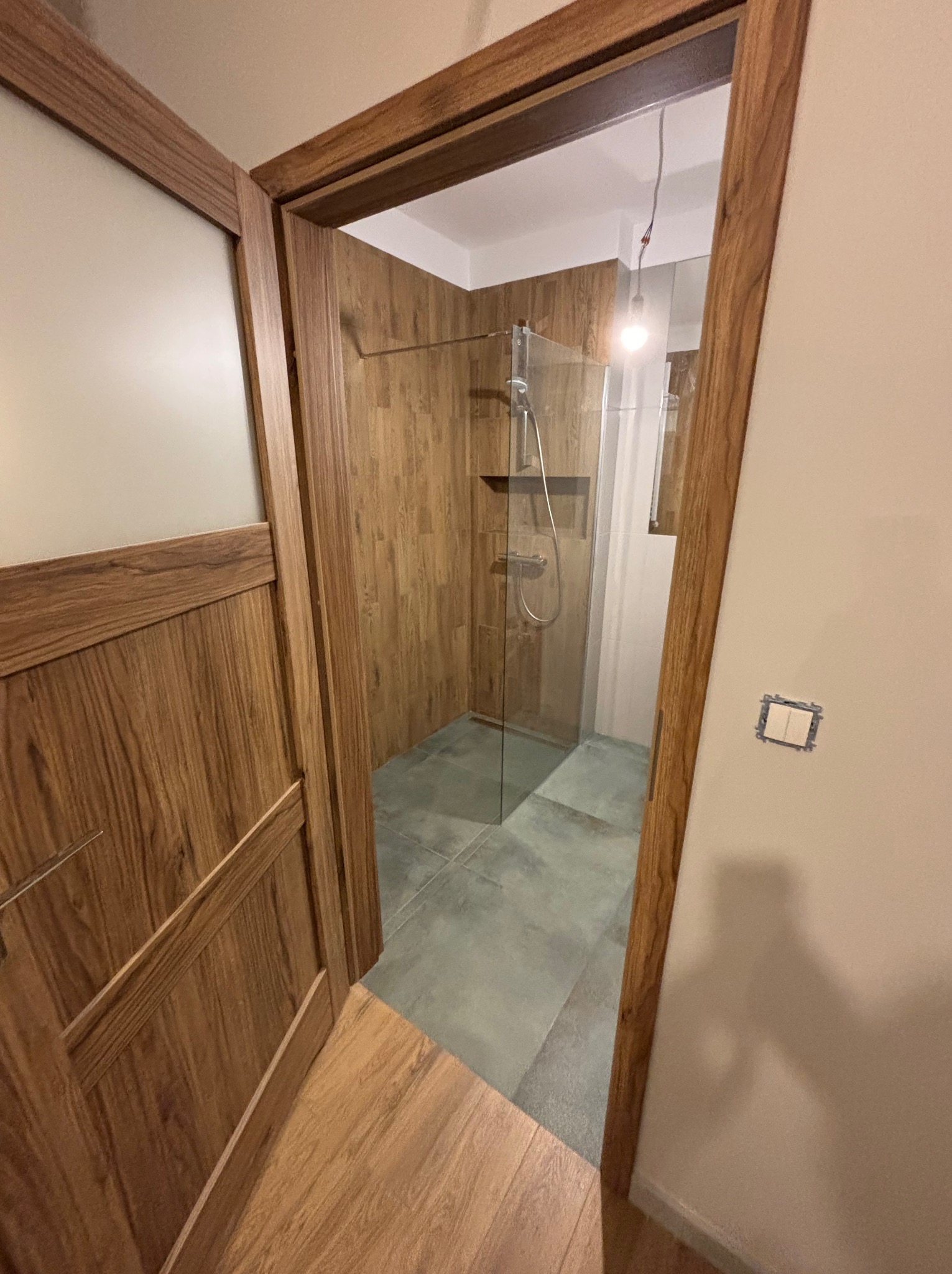 Drewniane wykończenie kabiny prysznicowej z szklaną ścianką, widok zza otwartych, drewnianych drzwi. Szare płytki na podłodze, minimalistyczny design.