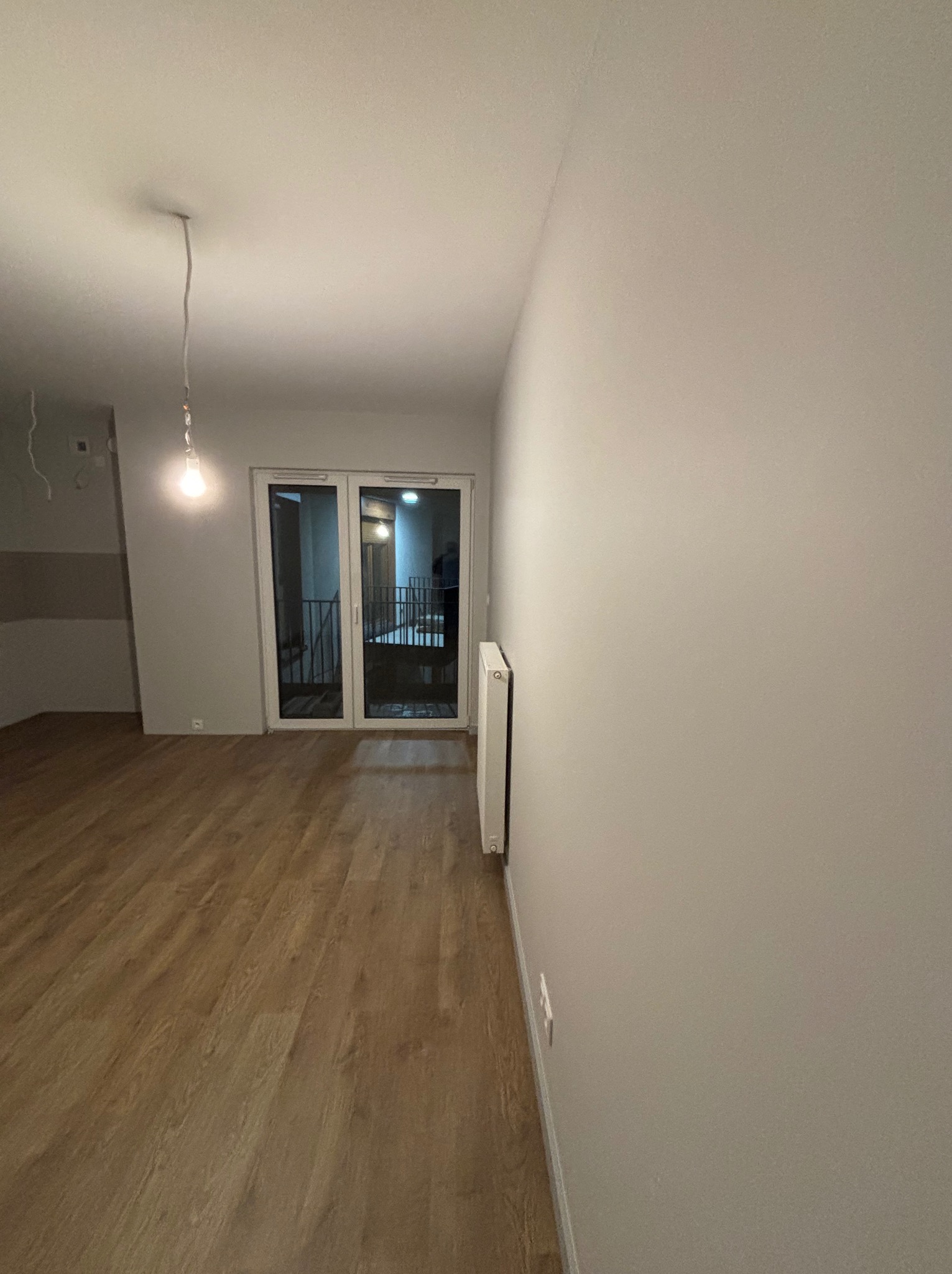 Pusty pokój z drewnianą podłogą i świeżo pomalowanymi ścianami na szaro. Widoczne drzwi balkonowe i wisząca żarówka na kablu. Minimalistyczne wnętrze przed umeblowaniem.