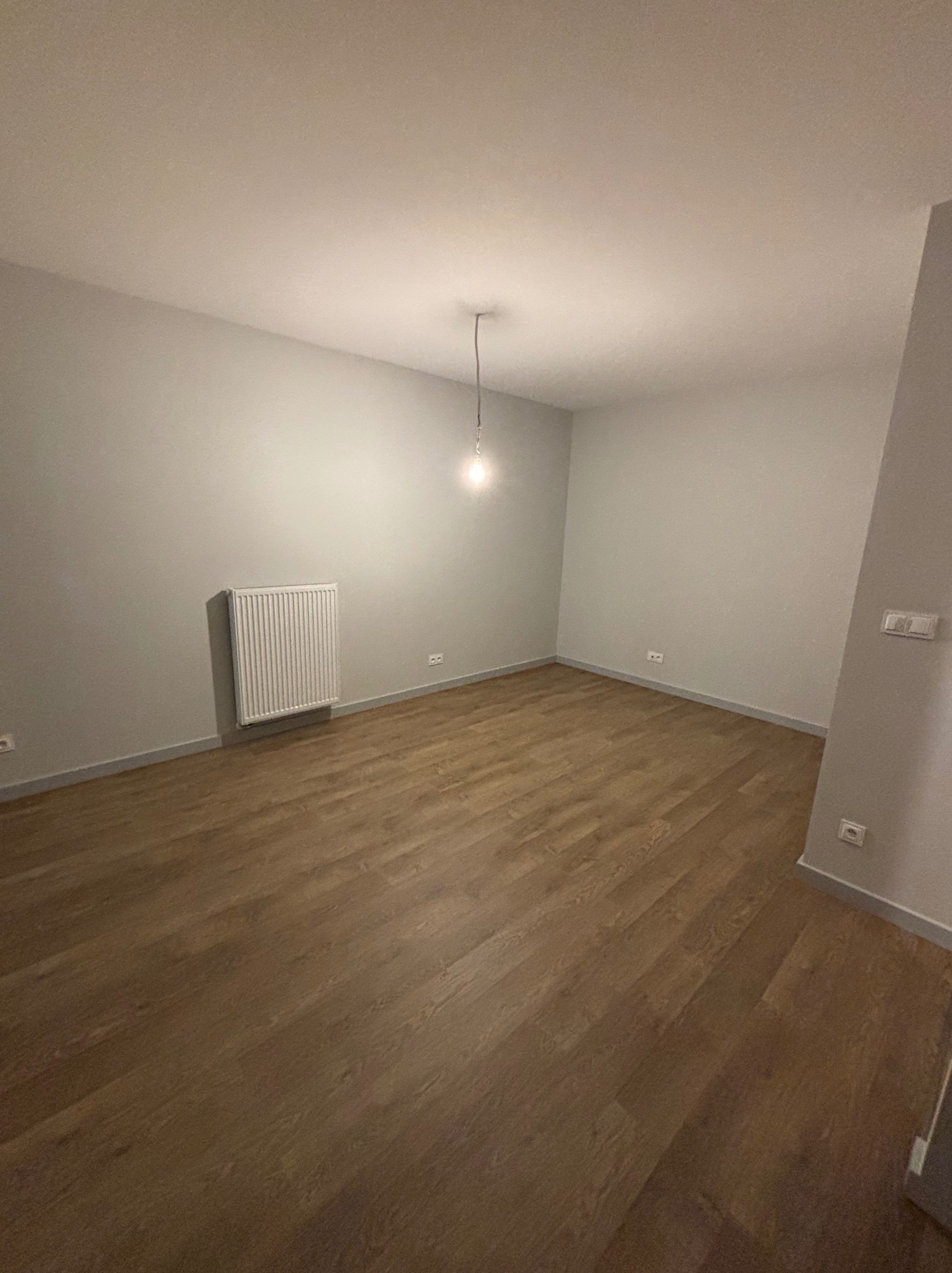 Jasne, puste pomieszczenie z drewnianą podłogą, szarymi ścianami i grzejnikiem. Minimalistyczne wnętrze, oświetlone pojedynczą żarówką.