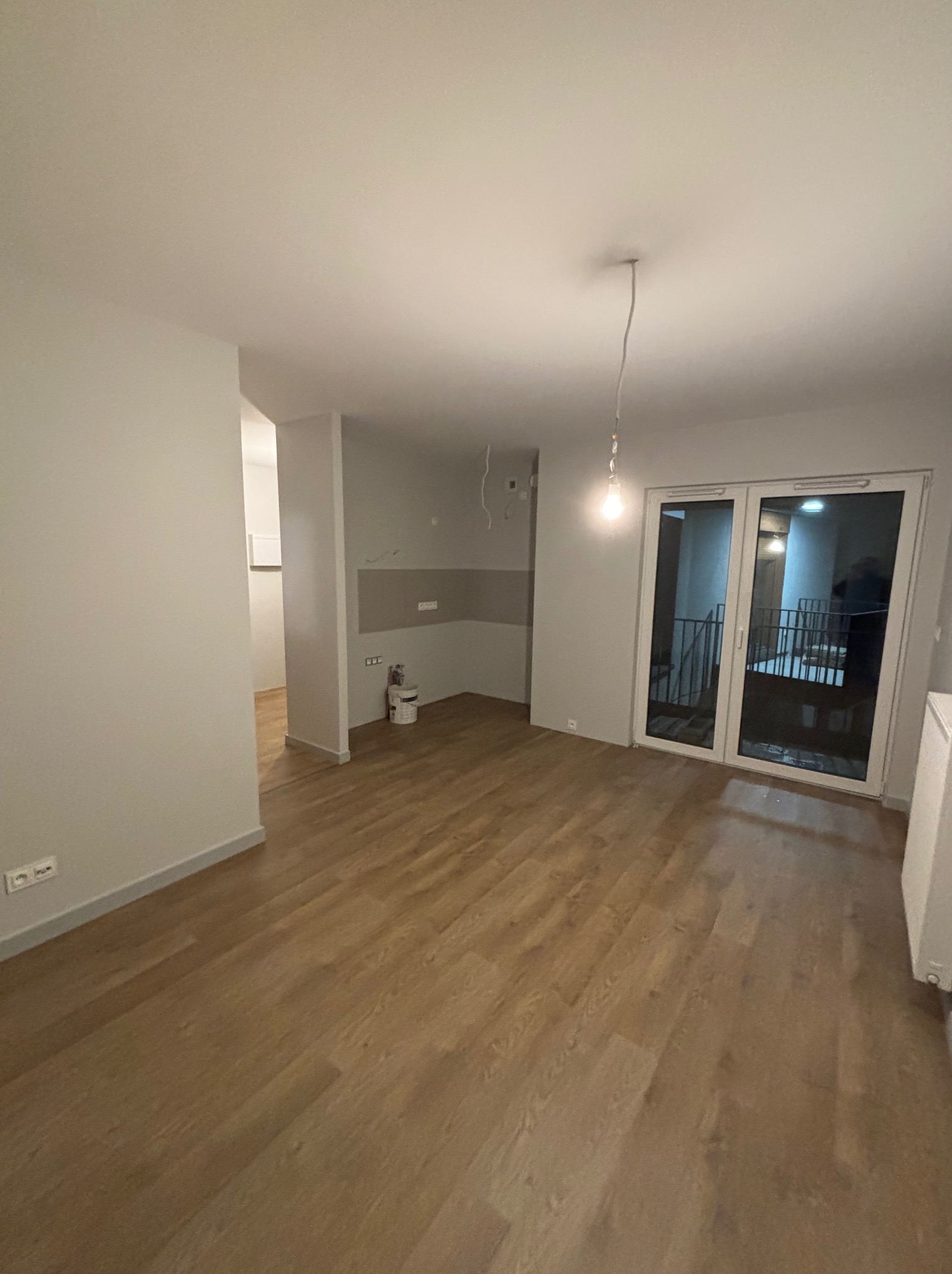 Jasne wnętrze po remoncie: parkiet, świeżo pomalowane ściany w odcieniach szarości, duże okno balkonowe. Widoczny fragment instalacji elektrycznej.