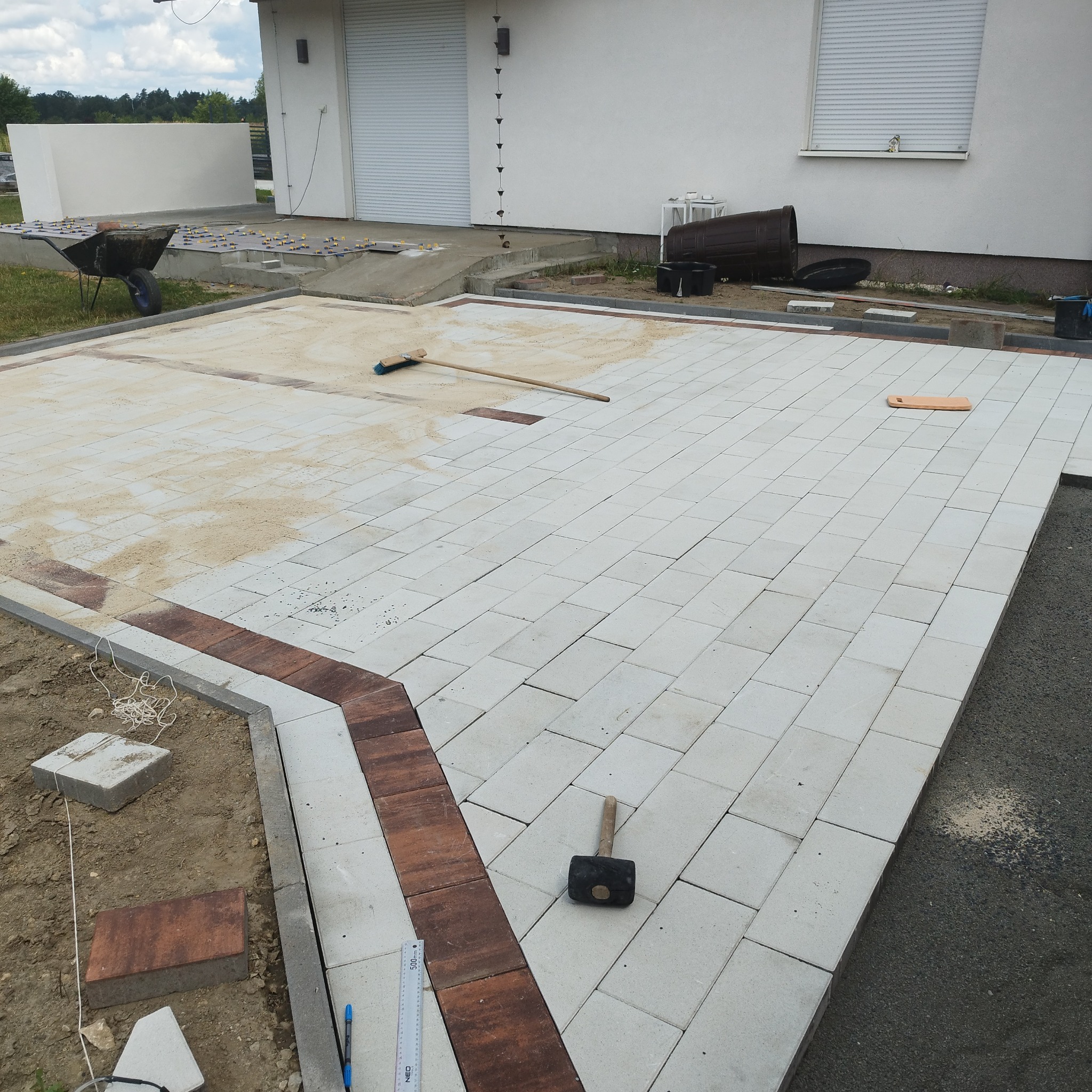 Częściowo ułożona, jasnoszara kostka brukowa z bordurą z ciemniejszej kostki. Widoczny młotek brukarski, miotła i taczka z materiałami. Etap prac na posesji.