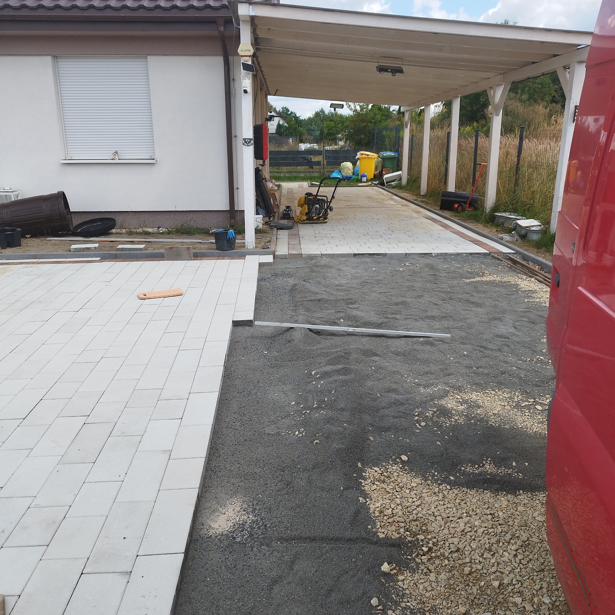 Fragment układania jasnej kostki brukowej na posesji. Część powierzchni wyłożona, obok przygotowane podłoże z piasku i kamienia. W tle budynek z podjazdem.